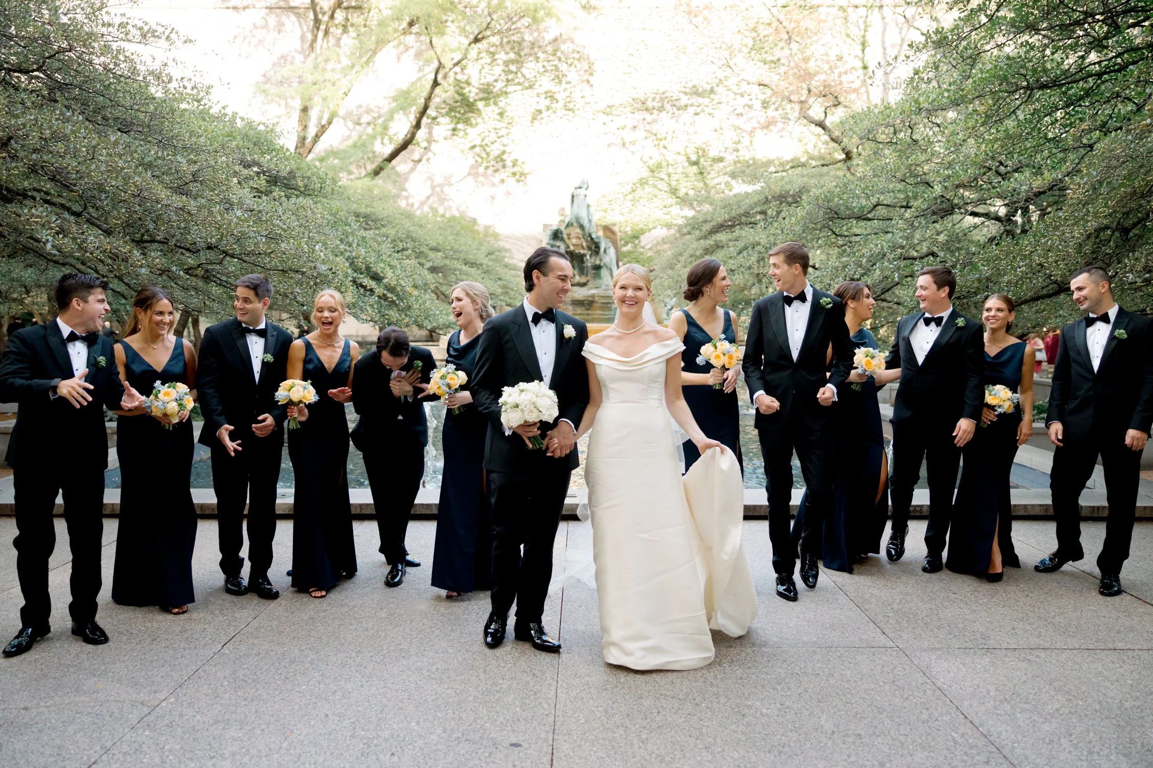 The full wedding party strolls before a stone garden fountain in black gowns and tuxedos, captured candidly on film by a Chicago film wedding photographer.