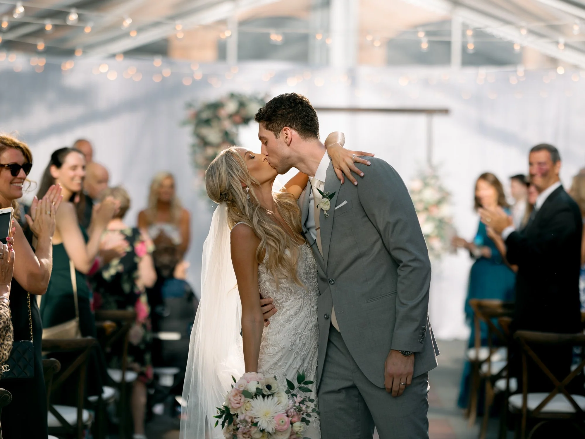 The bride and groom share their first kiss as a married couple under the glowing glass tent at the Chicago Illuminating Company, guests cheering all around.