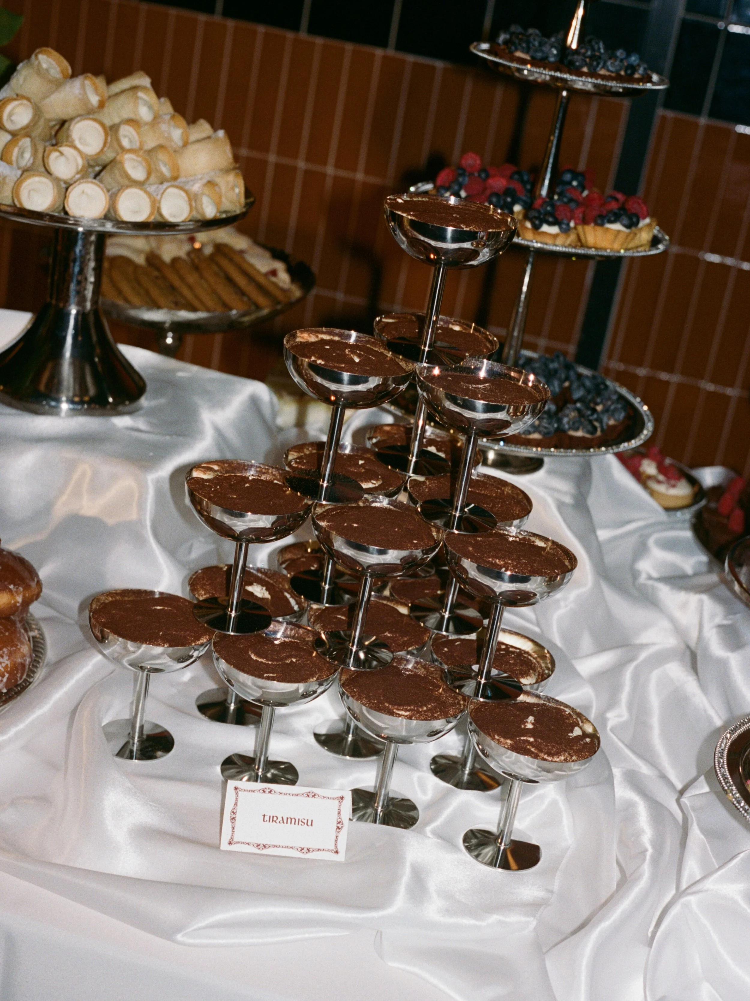 Tiramisu coupes stacked in a tower alongside cannoli and pastries on the dessert table