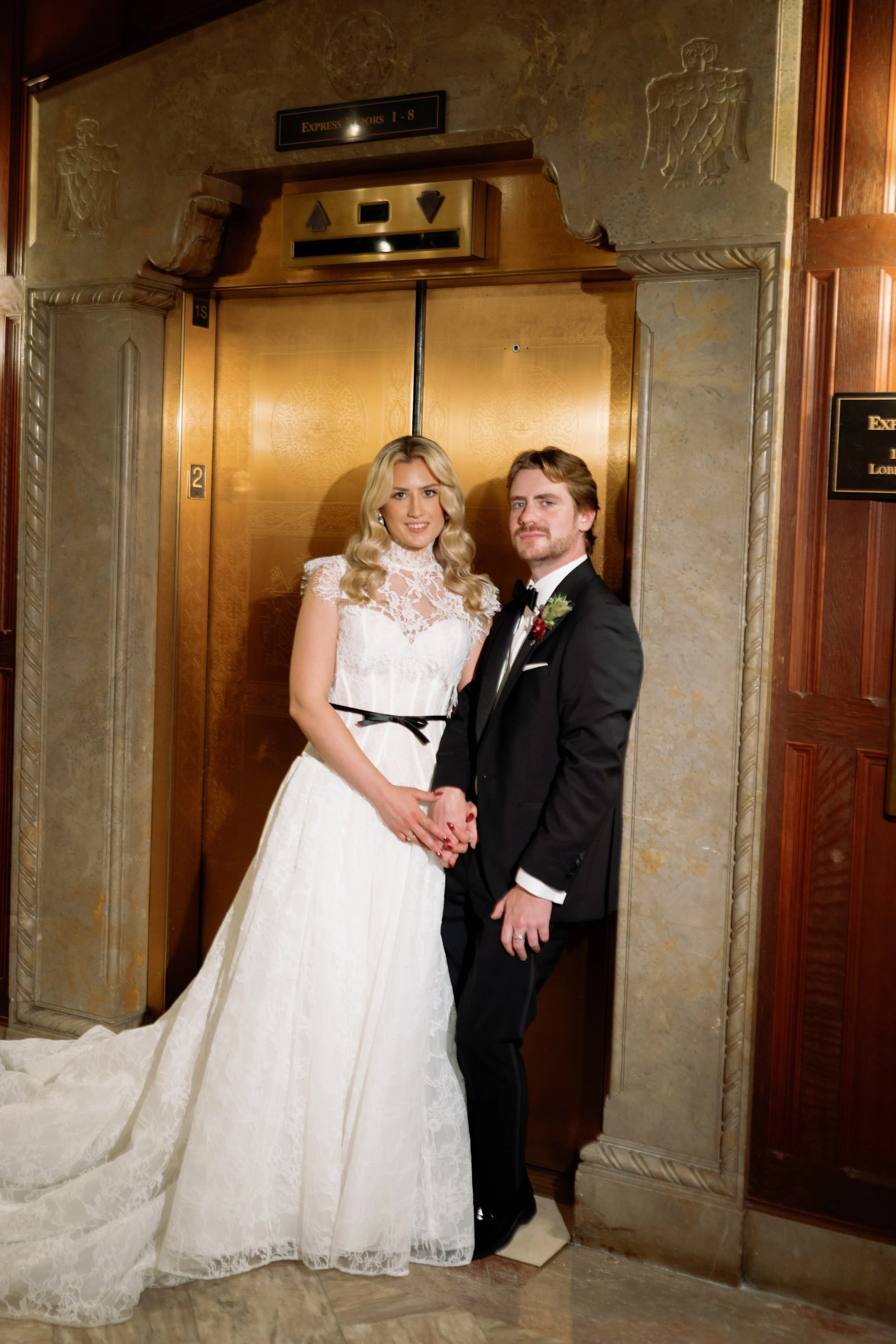Bride and groom hold hands in front of a vintage brass elevator in the hotel lobby