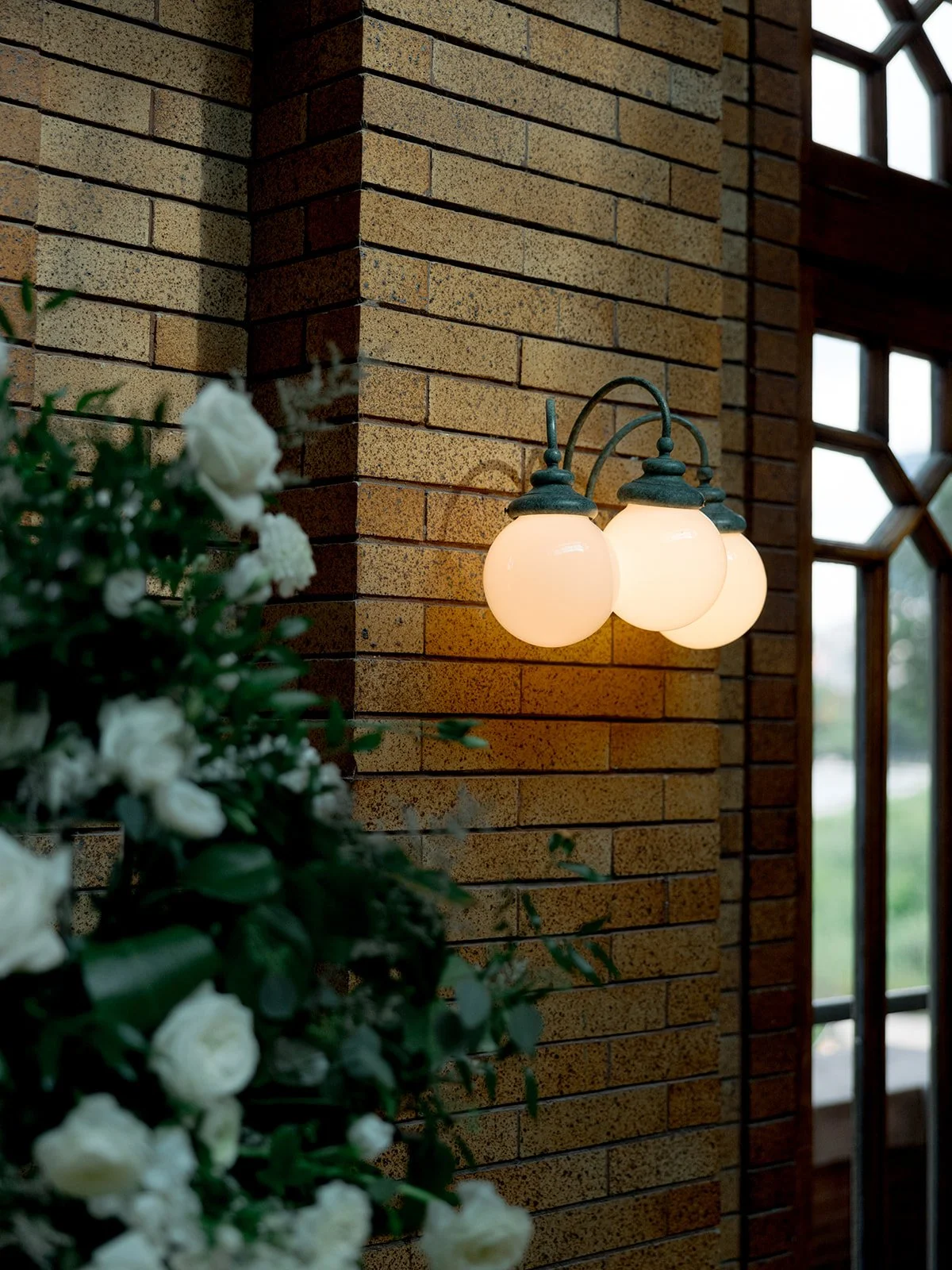 Warm globe wall sconce glowing against Cafe Brauer's tawny brick interior with a lush white rose and greenery arrangement.