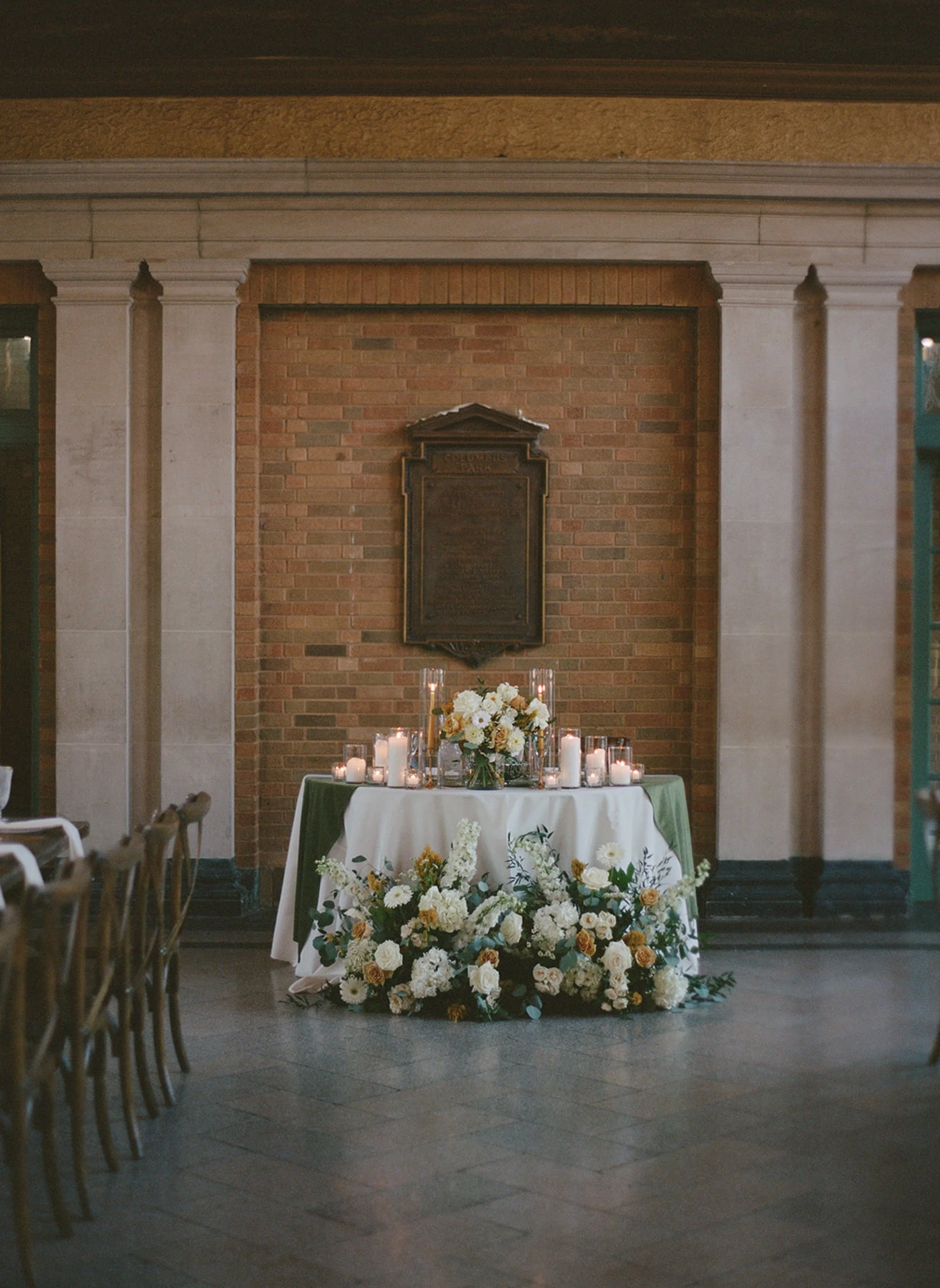 A voluminous white and golden floral arrangement cascades across the sweetheart table at Columbus Park Refectory — pillar candles glowing, the historic brick wall and ornate plaque as backdrop