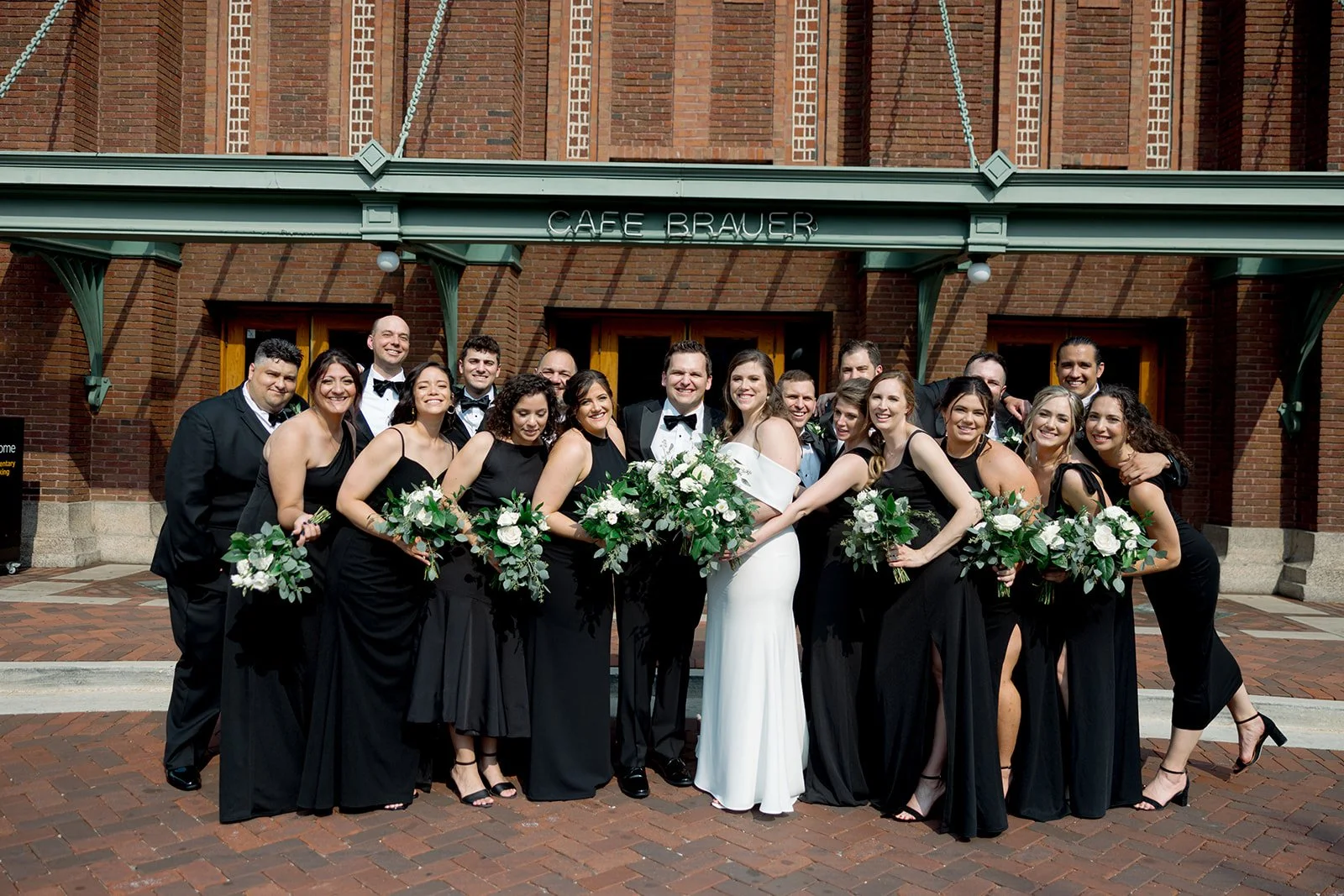 Full wedding party crowded together in front of the Cafe Brauer entrance — tuxedos, black gowns, bride in white at center.