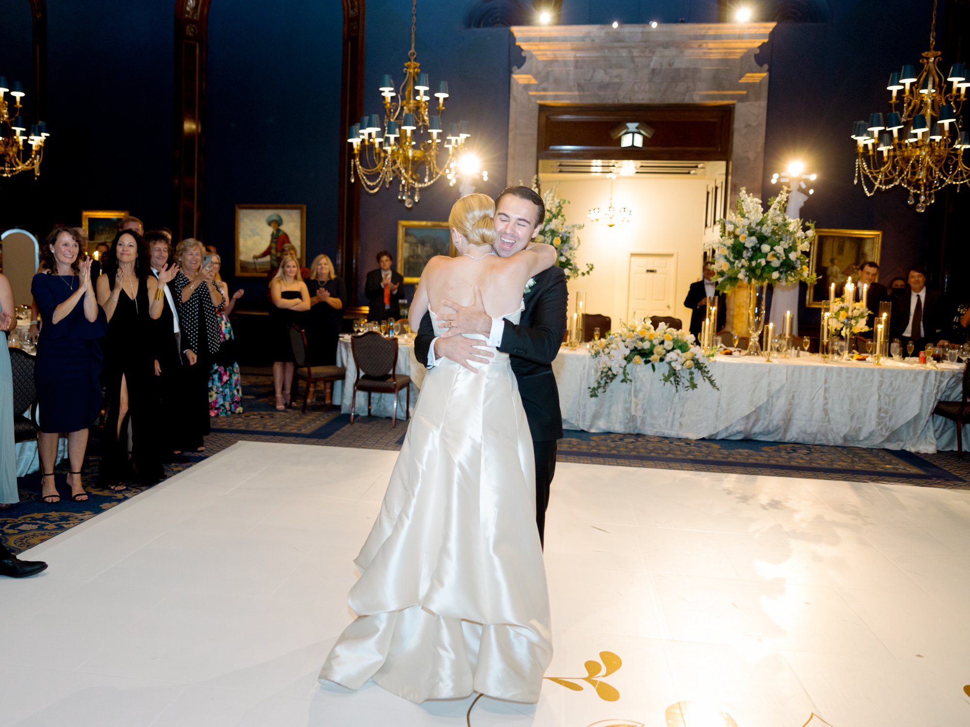 The groom pulls the bride into a full embrace mid-first dance, his smile filling the frame while guests cheer in the navy Union League Club ballroom.
