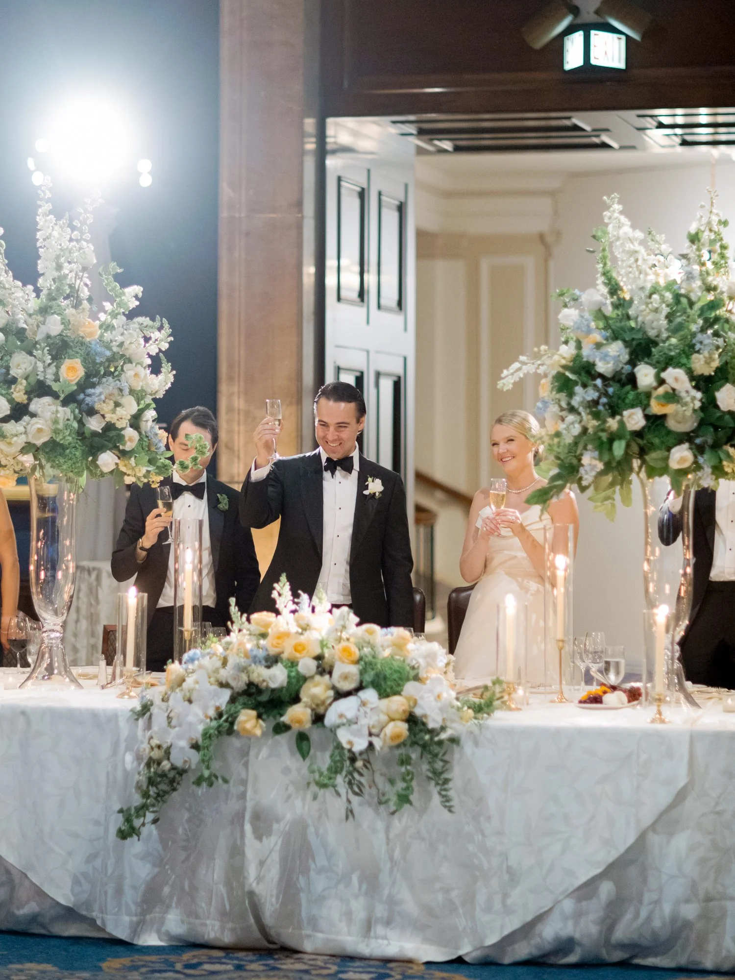 The groom lifts his champagne flute at the sweetheart table, flanked by towering white and yellow floral arrangements in the Union League Club ballroom.