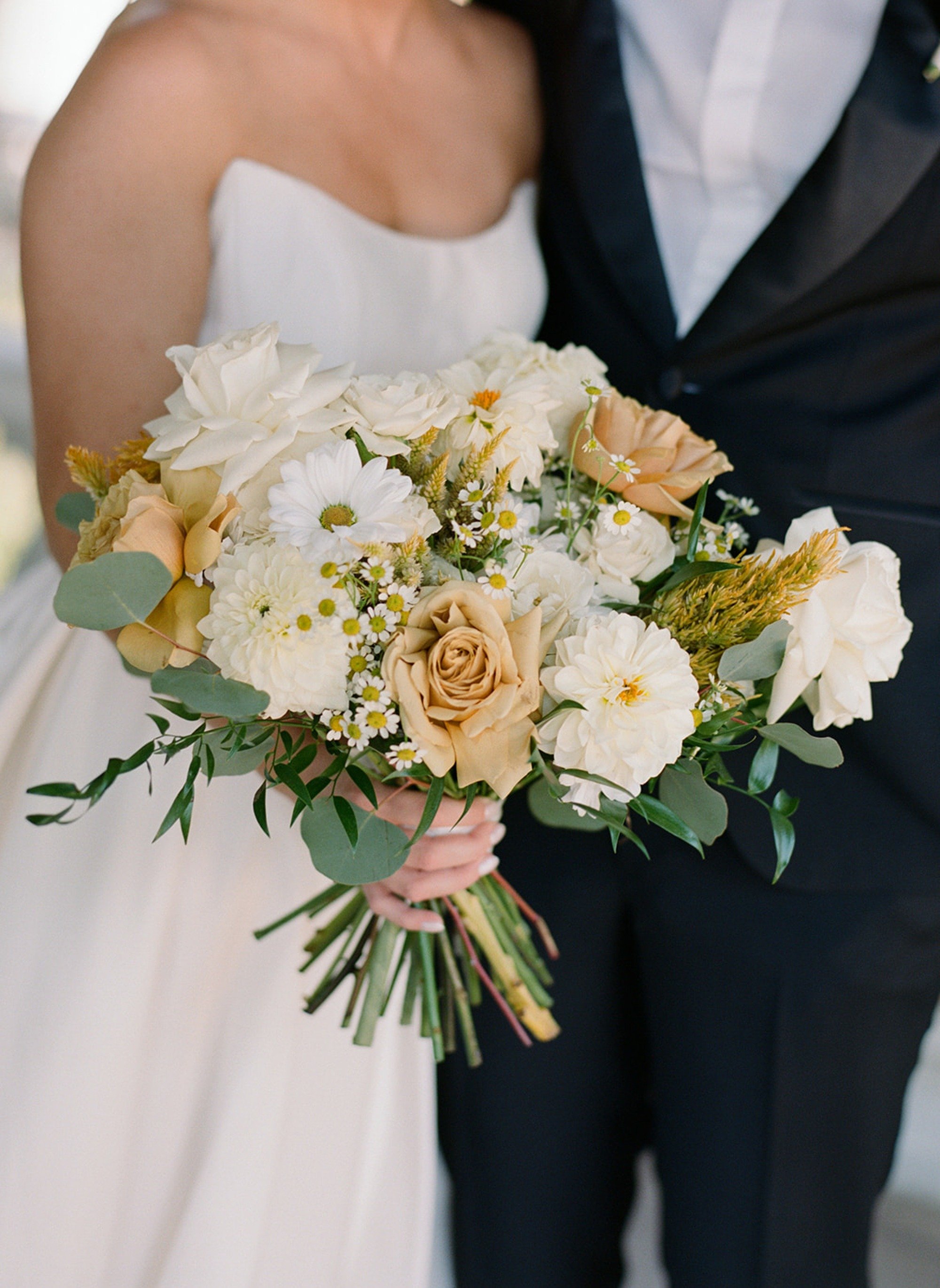 A lush bridal bouquet close-up — champagne garden roses, white dahlias, daisies, and eucalyptus gathered loosely against the bride's strapless gown. Wild, romantic, and perfectly imperfect. Columbus Park Refectory