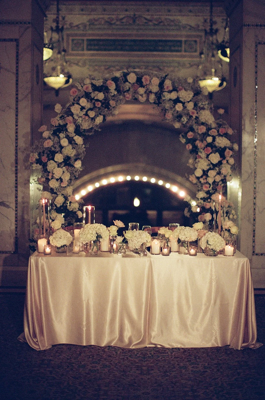 The sweetheart table beautifully set beneath an arched white floral installation at Chicago Cultural Center. Elegant candlelight and lush blooms create an intimate reception moment. Shot on film by a Chicago film wedding photographer.