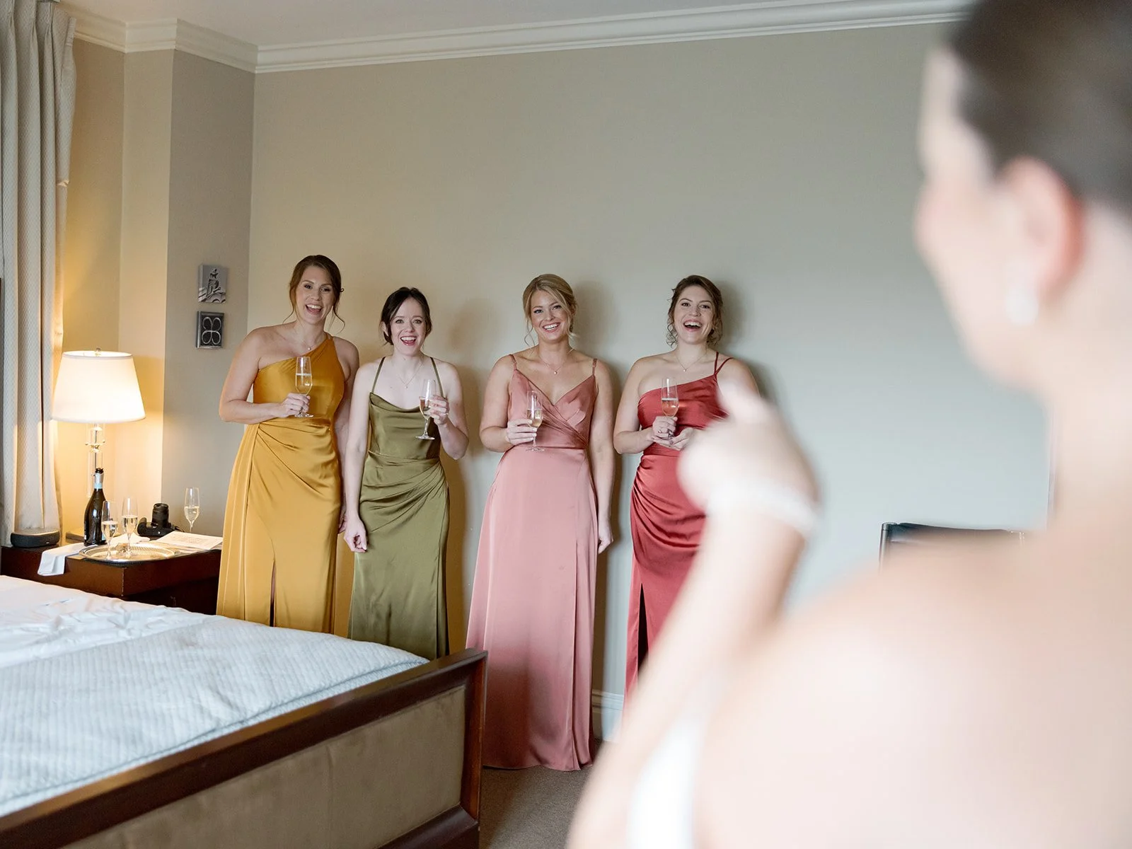 Four bridesmaids in rust, gold, pink, and olive gowns react with delight as the bride walks in — their faces say everything about how she looks. A getting-ready moment full of genuine emotion — captured by a Chicago film wedding photographer.