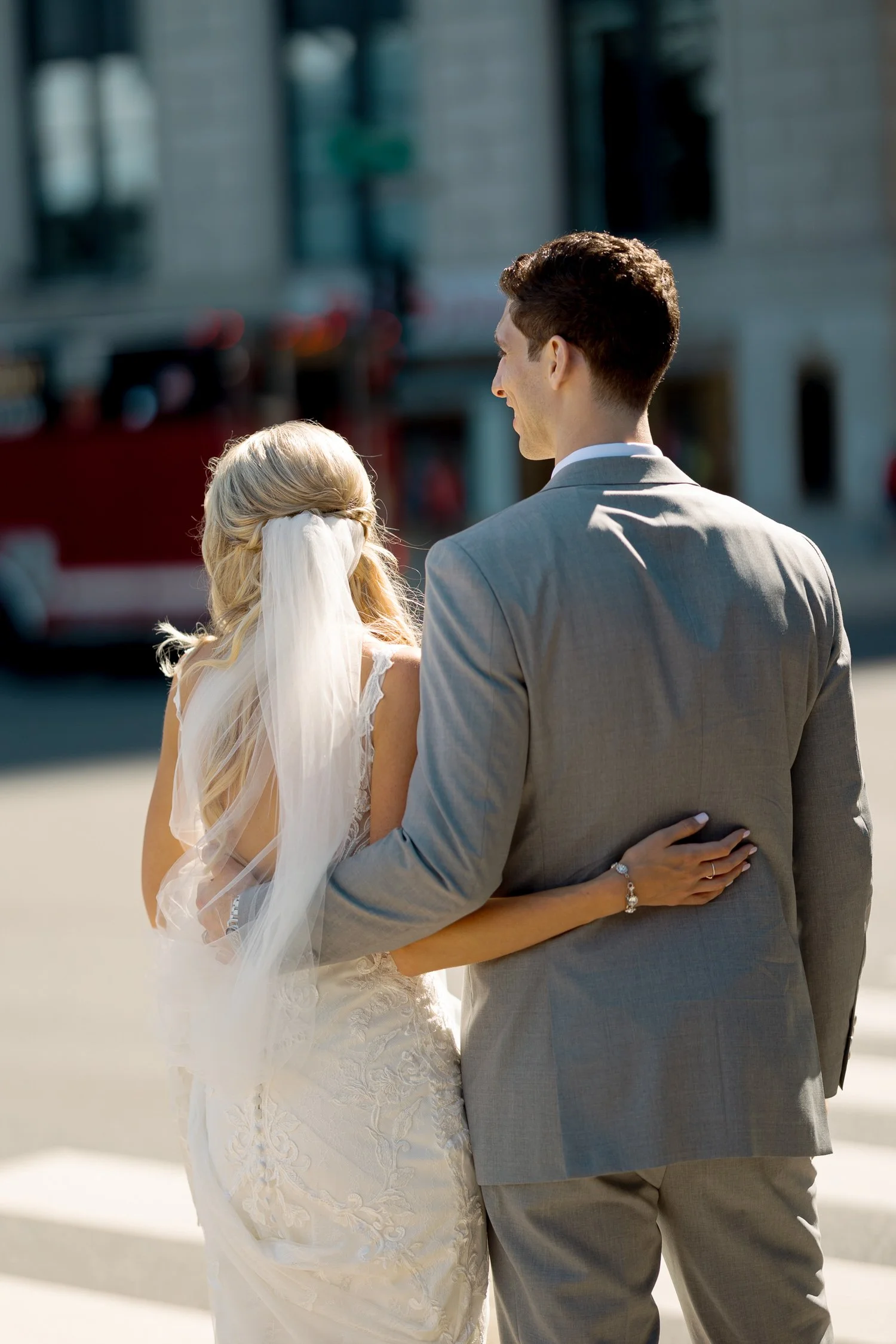 The bride and groom walk arm-in-arm through downtown Chicago streets, seen from behind as the city buzzes around them on their wedding afternoon.