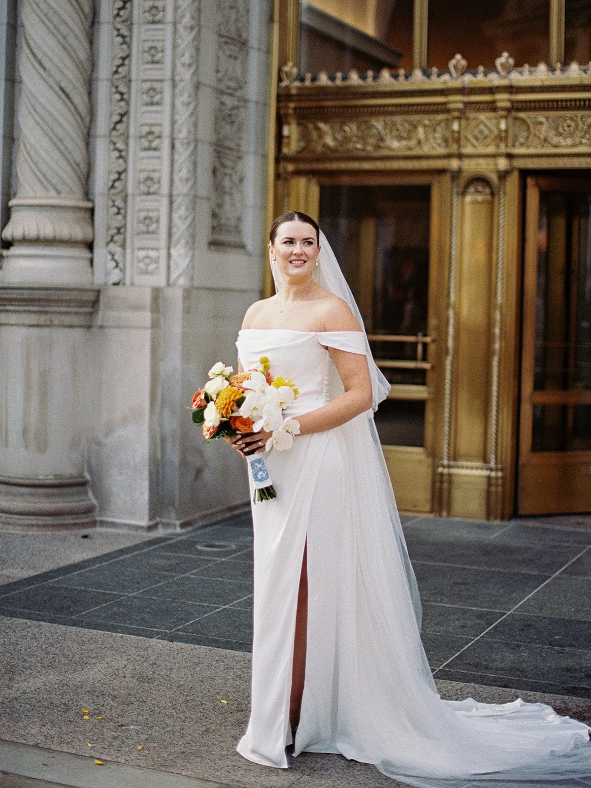 The bride stands in front of a gold-detailed Chicago building entrance — gown slit open, cathedral veil trailing, bouquet in hand and a smile that commands the whole frame.