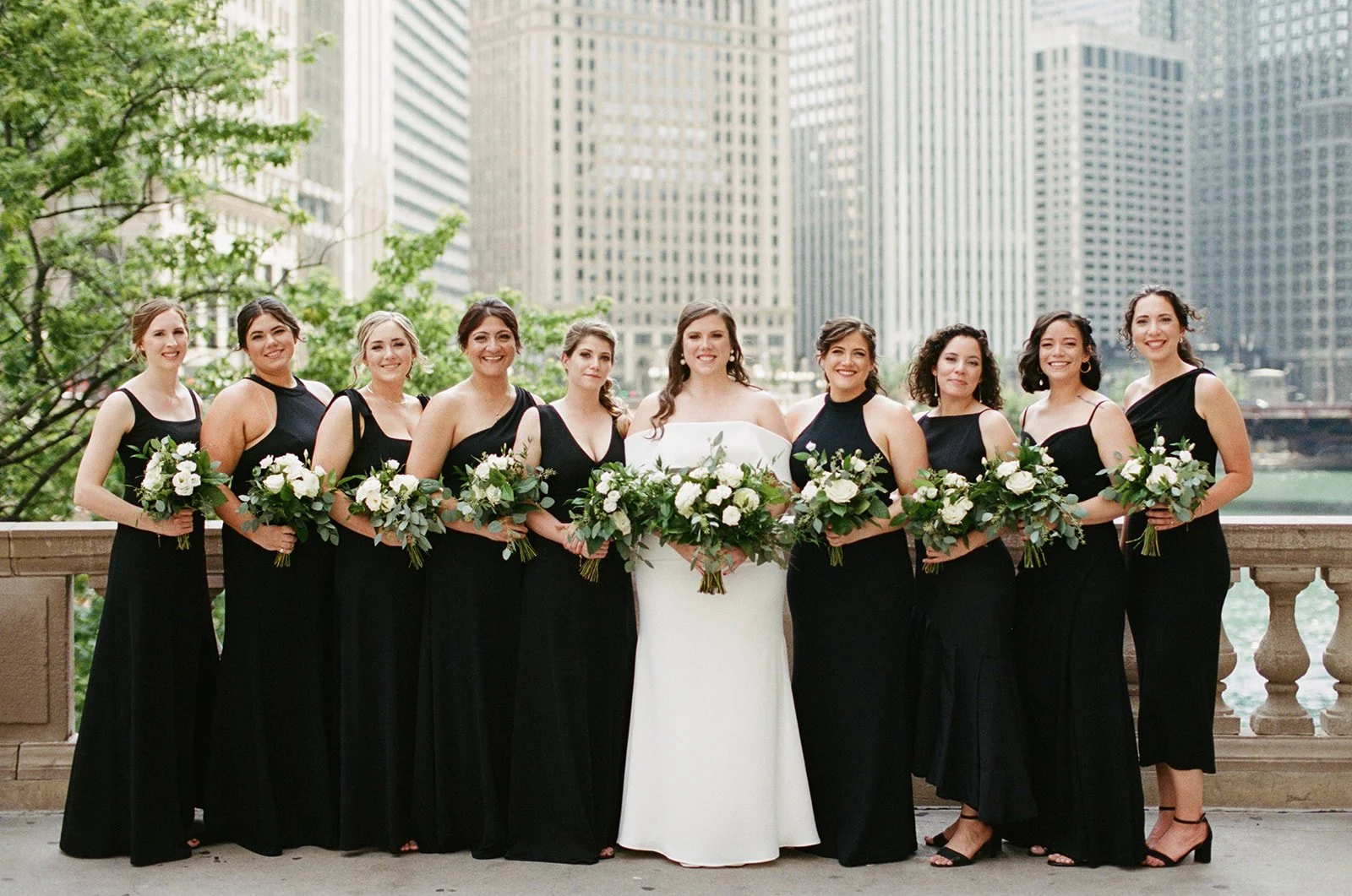 Nine bridesmaids in elegant black gowns flanking the bride on a stone terrace overlooking the Chicago River and skyline.
