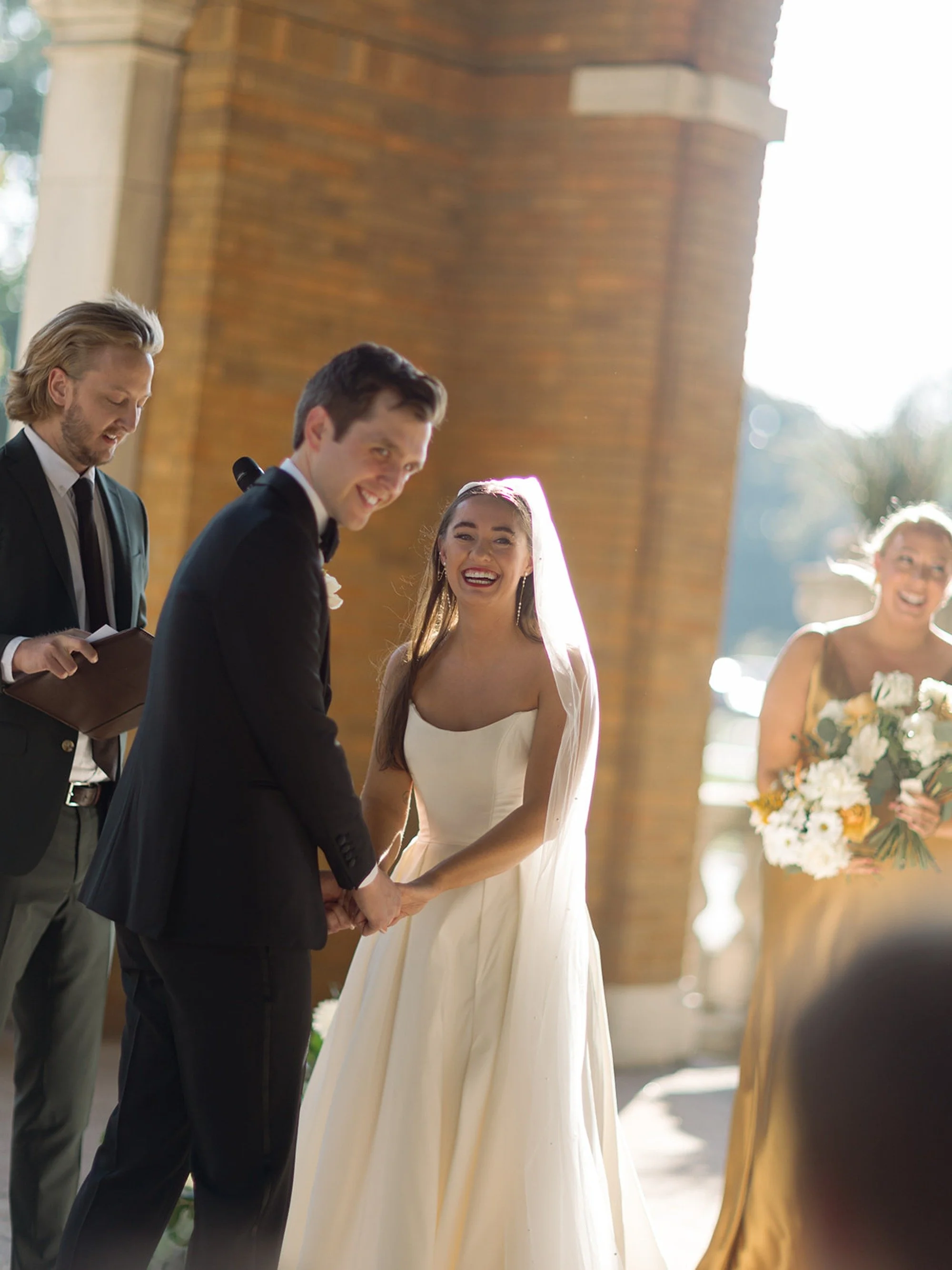 The bride beams — genuinely, completely, radiantly — during the ceremony at Columbus Park Refectory, golden afternoon sunlight flooding in and a maid of honor smiling just behind her. Pure joy, captured on film