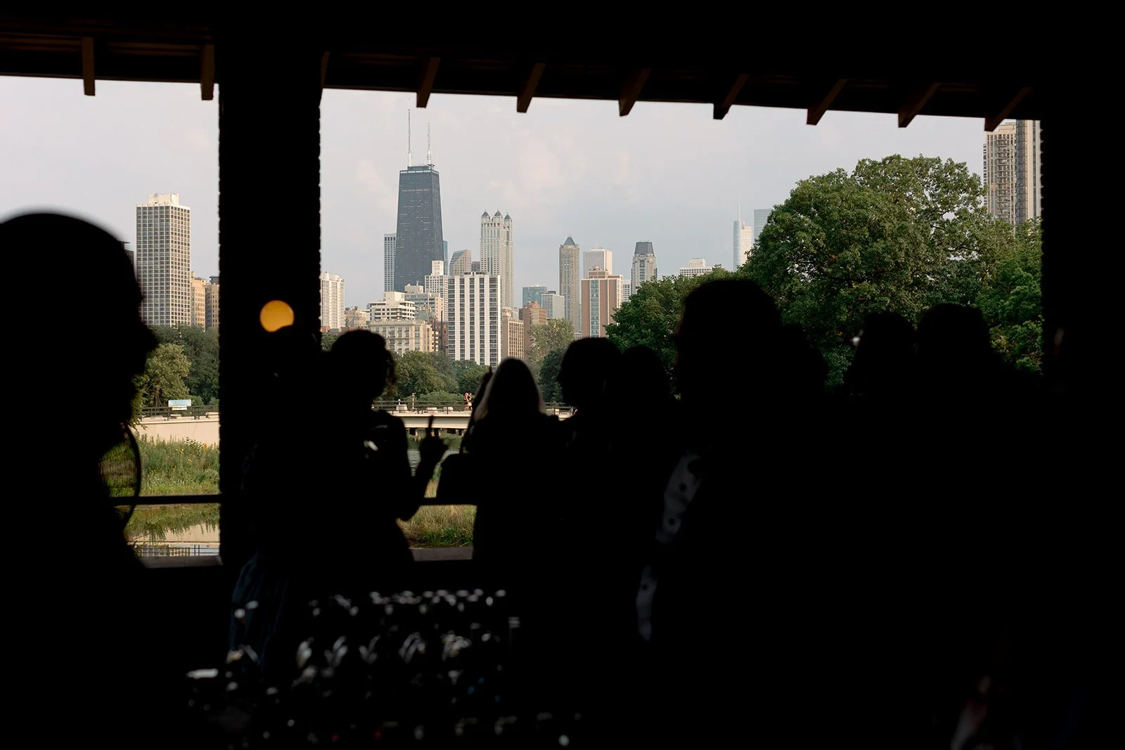 Guests silhouetted on the Cafe Brauer terrace at cocktail hour, the Chicago skyline and Lincoln Park lagoon glowing behind them.