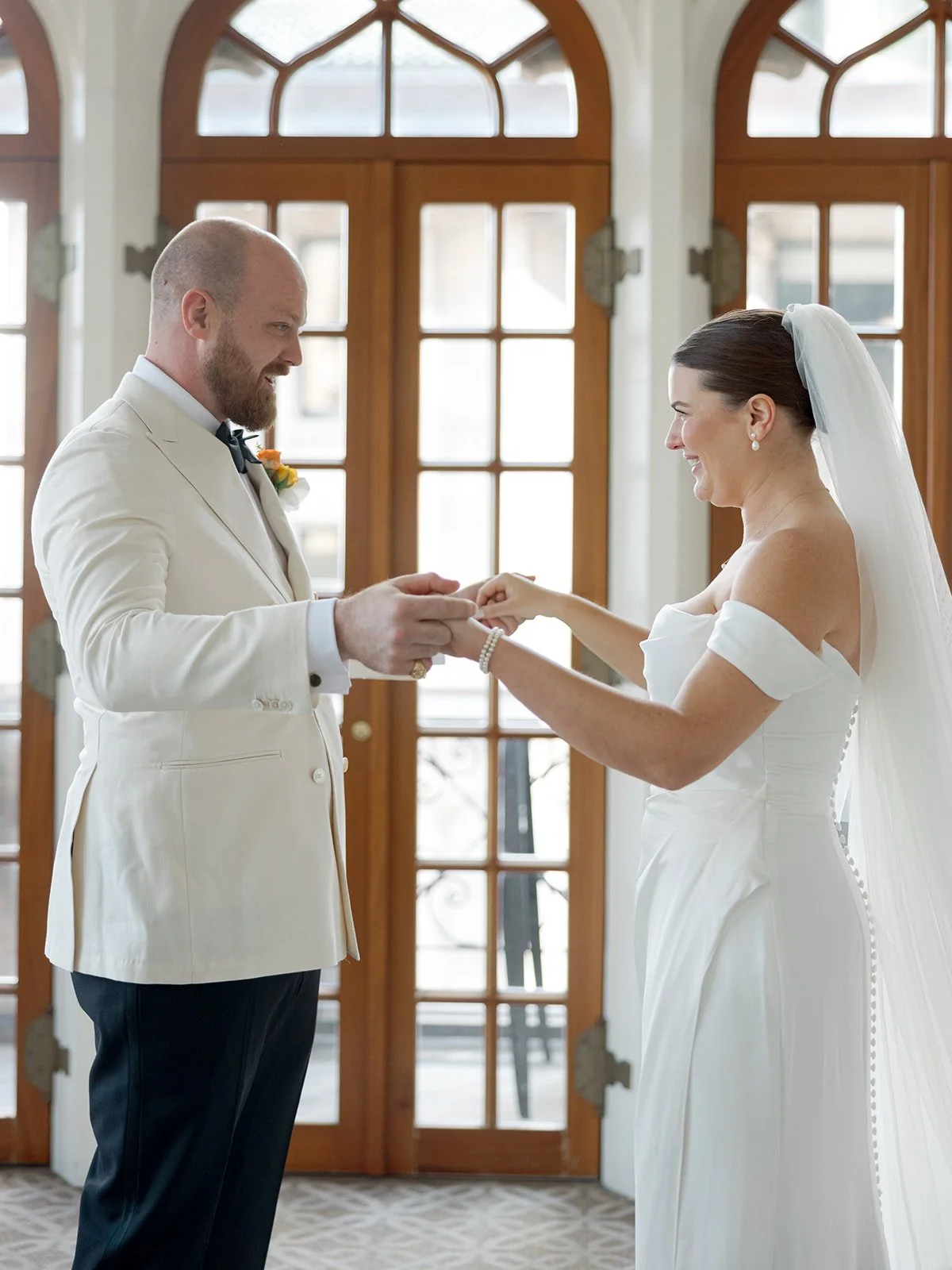 The first look ring exchange — the groom slides the band onto her finger and she breaks into a full, genuine laugh, French doors glowing warmly behind them. A moment of pure, unguarded joy