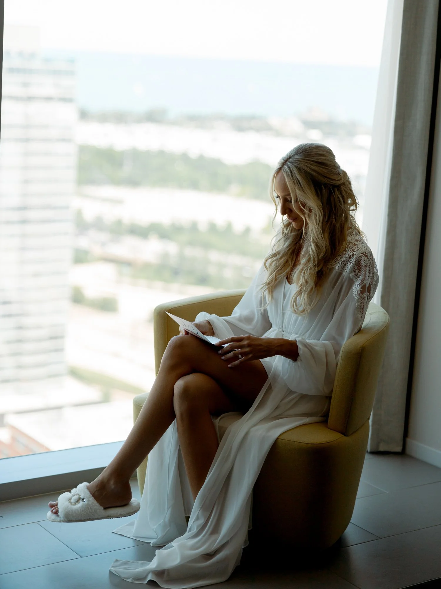 The bride sits in a hotel armchair reading a heartfelt letter on her wedding morning, the vast city view stretching behind her through the floor-to-ceiling window.