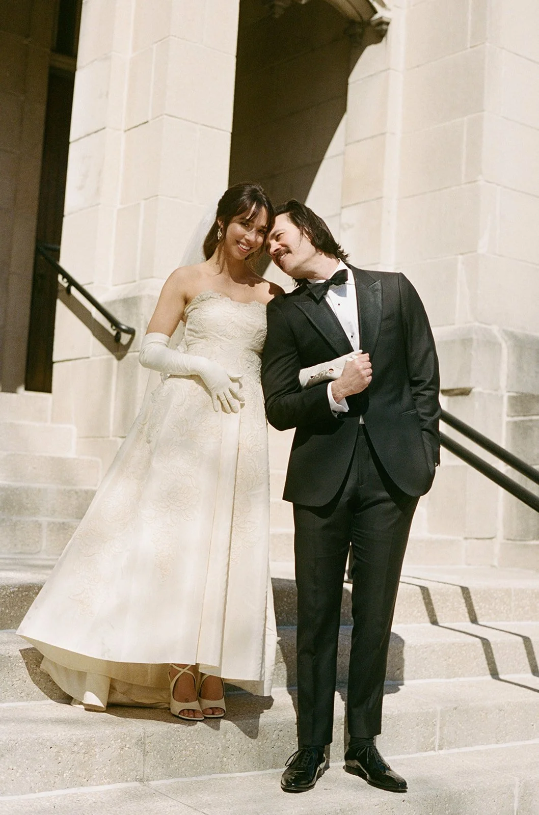 Film portrait of a bride in a lace ball gown with opera gloves and groom in a black tuxedo laughing together on stone steps at Barrington's White House, photographed by Louie Abellera.