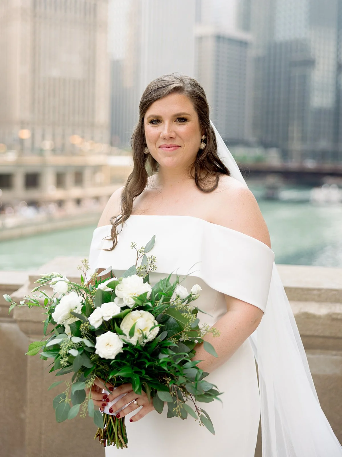 Close bridal portrait on the Chicago Riverwalk — the bride holding her white bouquet, city skyline shimmering behind her.