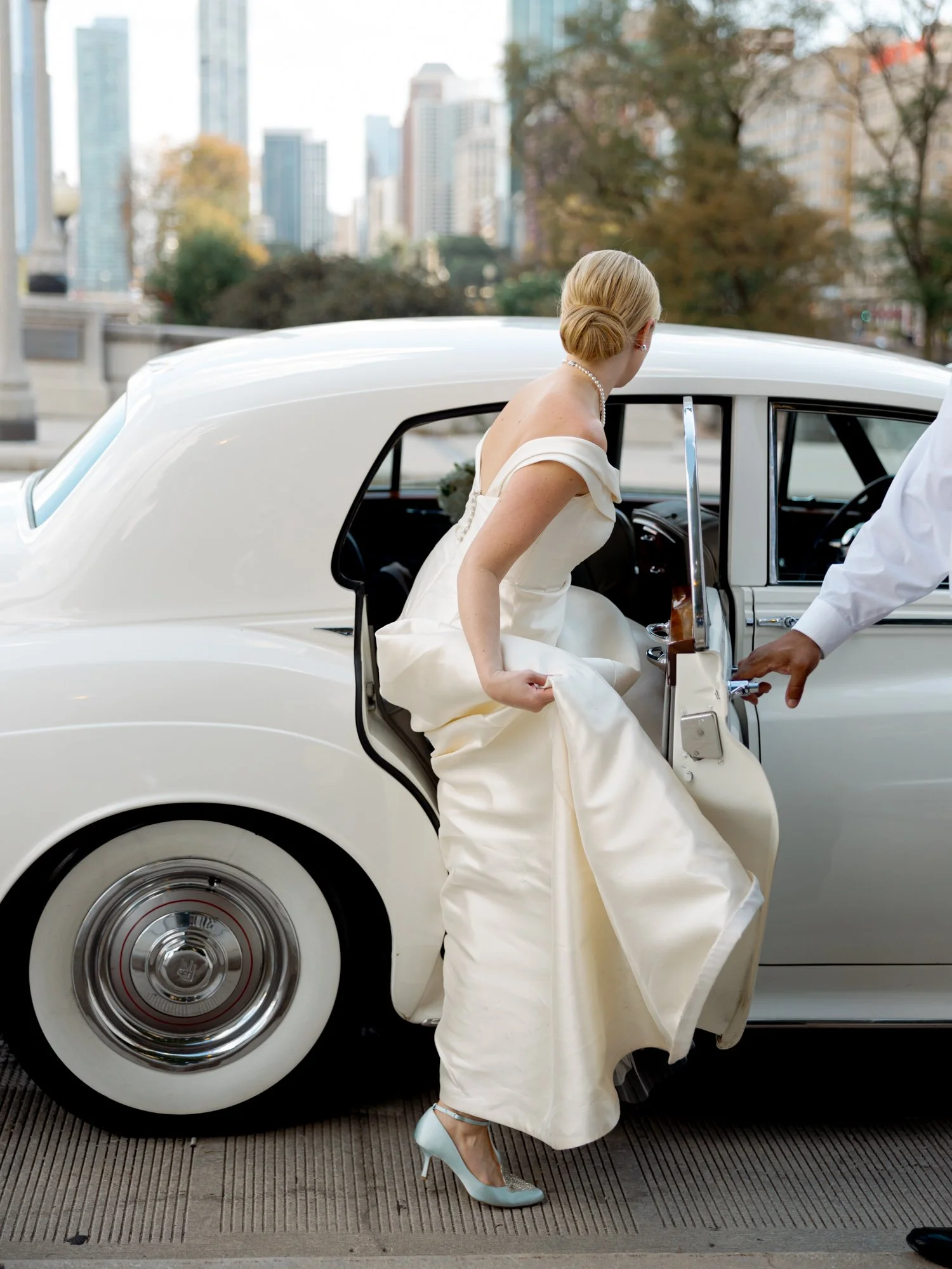 The bride lifts her silk gown train and steps into a vintage white Rolls-Royce, the full Chicago skyline rising dramatically behind her in the frame.