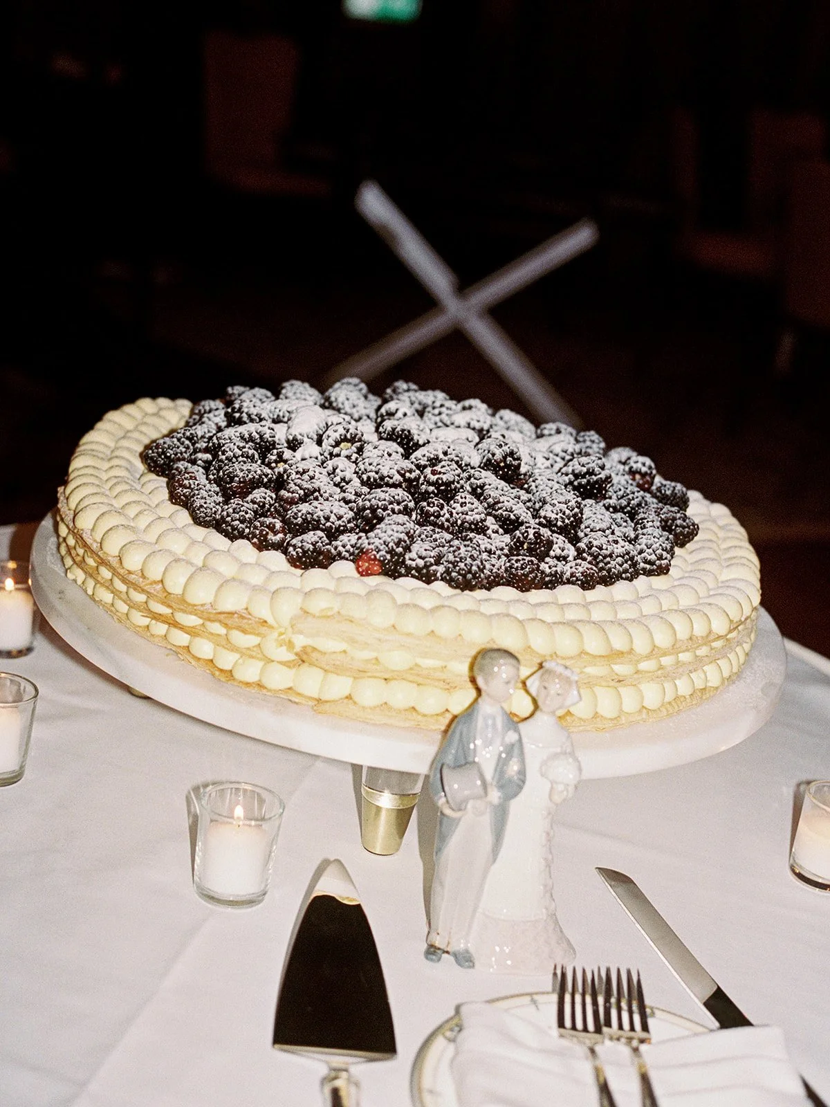 The wedding cake: an oval white cake topped with a mountain of fresh blackberries dusted in powdered sugar, a vintage ceramic bride-and-groom topper nestled at its base. Classic meets unexpected — captured by a Chicago film wedding photographer.