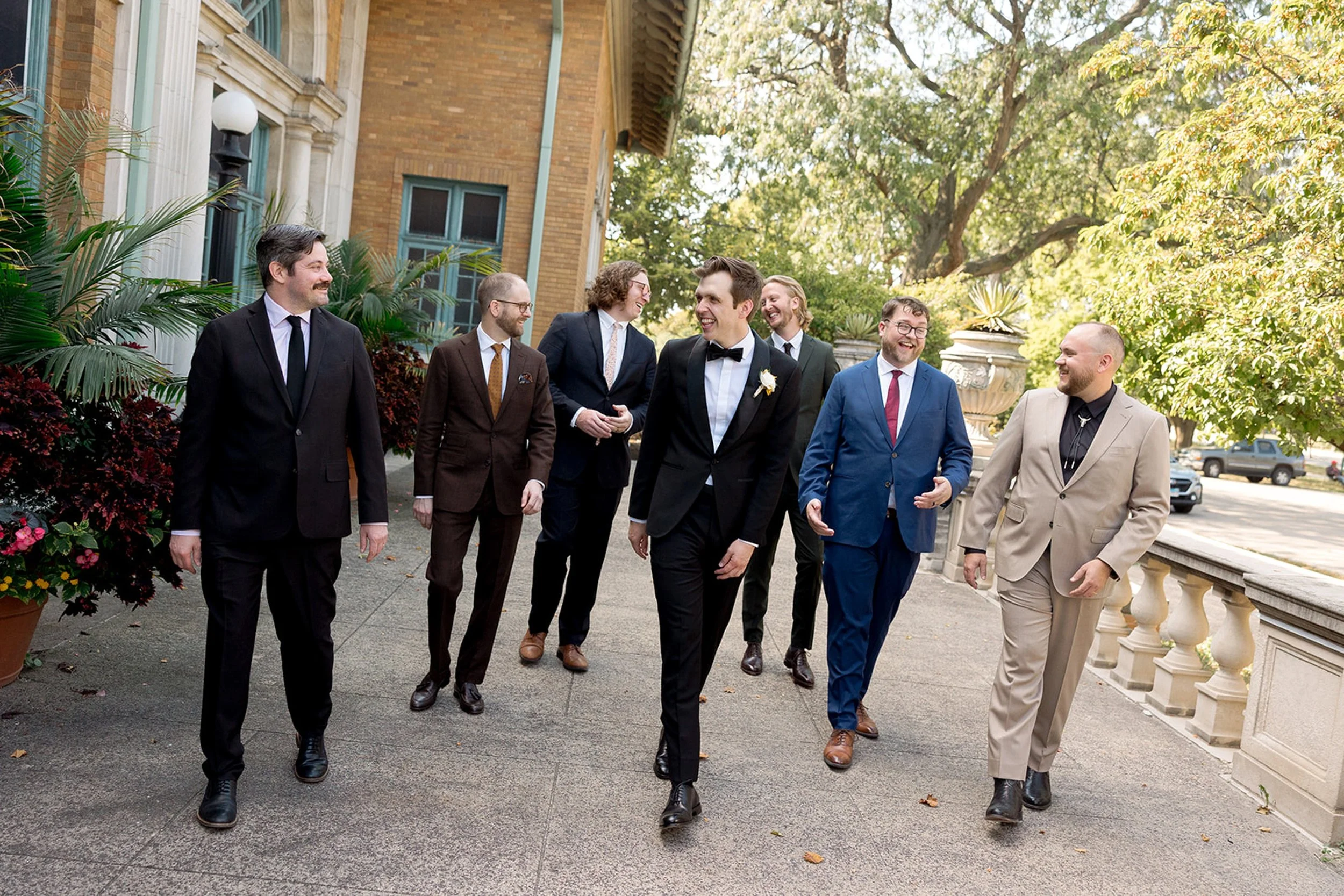 The groom and his groomsmen walk together in front of Columbus Park Refectory — a candid, easy portrait of sharp suits and genuine laughter. The groom in the center, right where he belongs