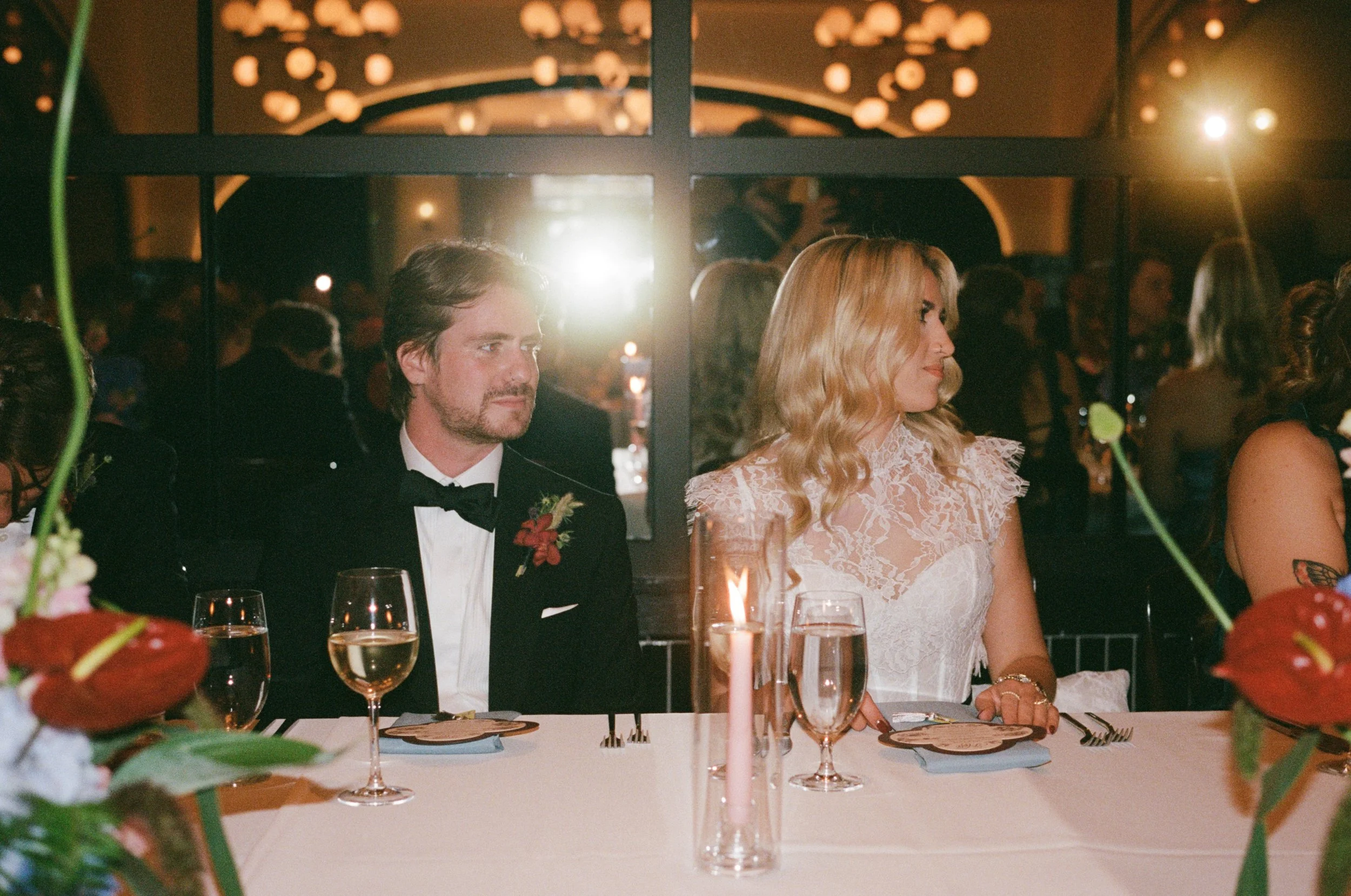 Bride and groom sit together at their candlelit reception table with guests nearby