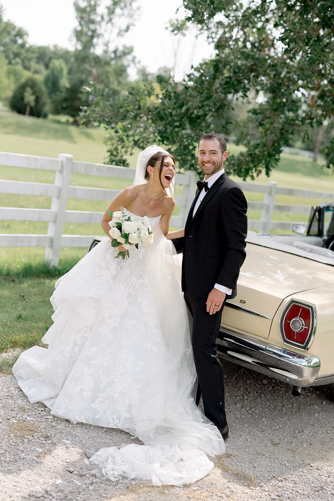 Candid laughing bride in a lace ball gown and groom in a black tuxedo leaning against a vintage car at a private estate wedding, photographed by Louie Abellera.