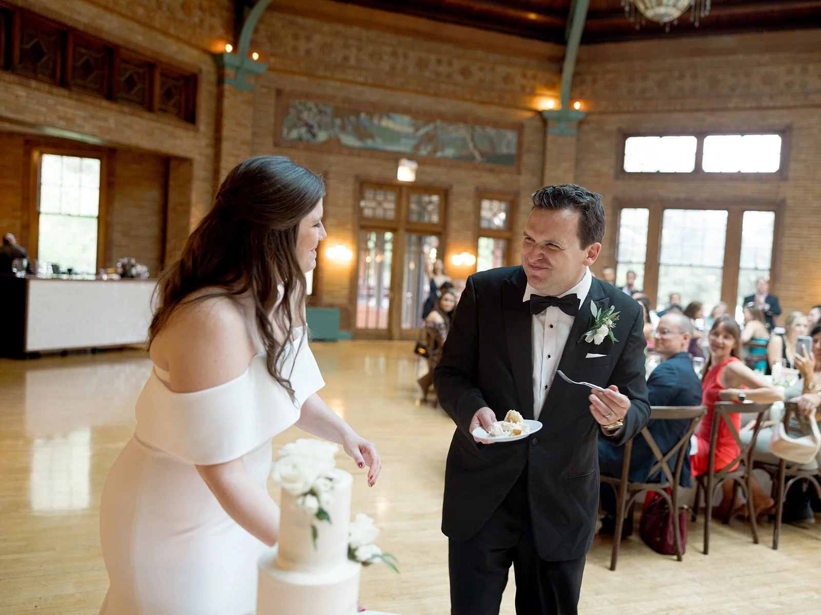 Groom making a comically suspicious face as the bride holds up a slice of wedding cake at Cafe Brauer's reception.