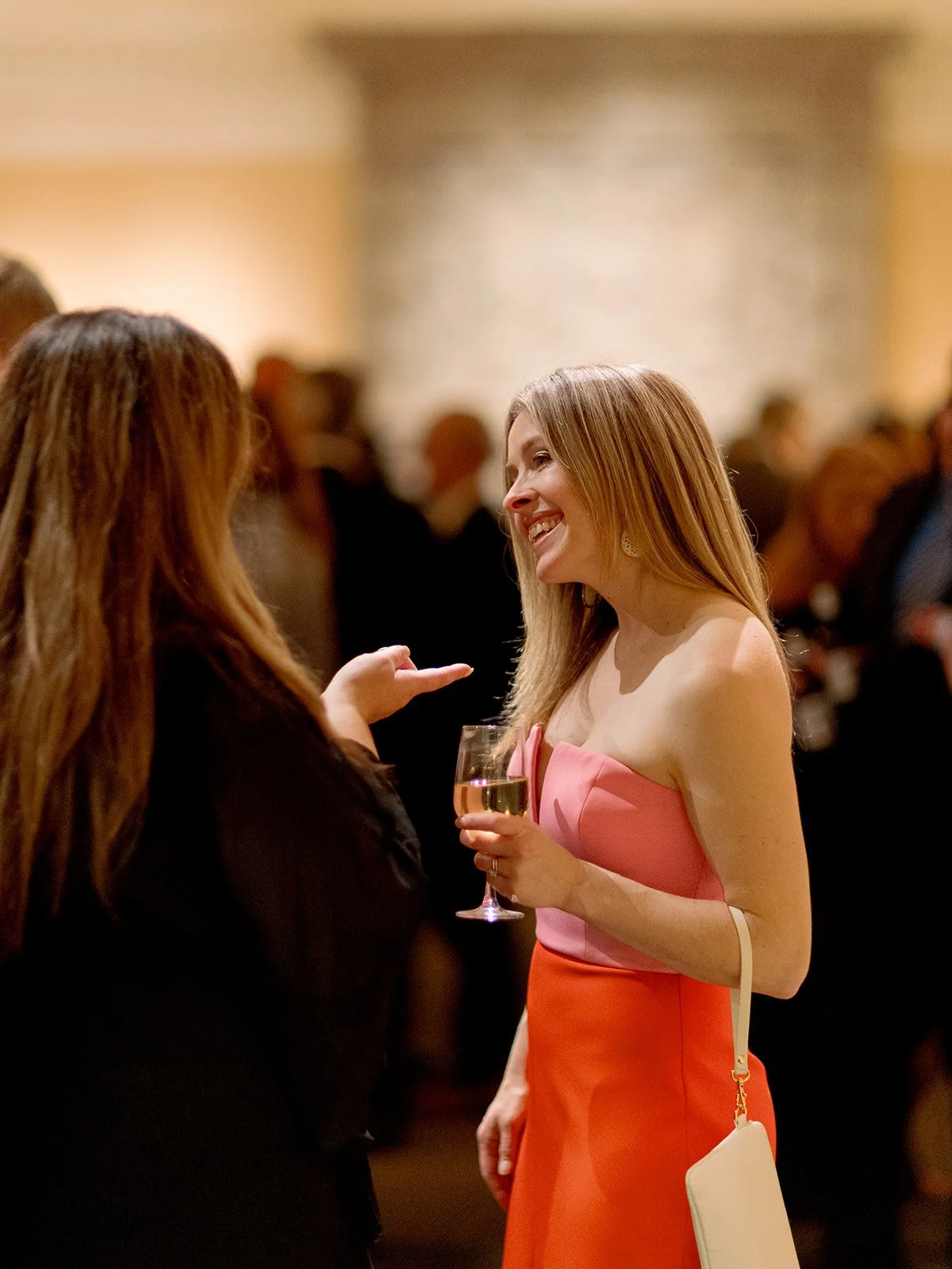 A guest in a vibrant pink-to-orange dress laughs in the middle of a conversation during cocktail hour, champagne glass raised, the room buzzing with energy behind her.