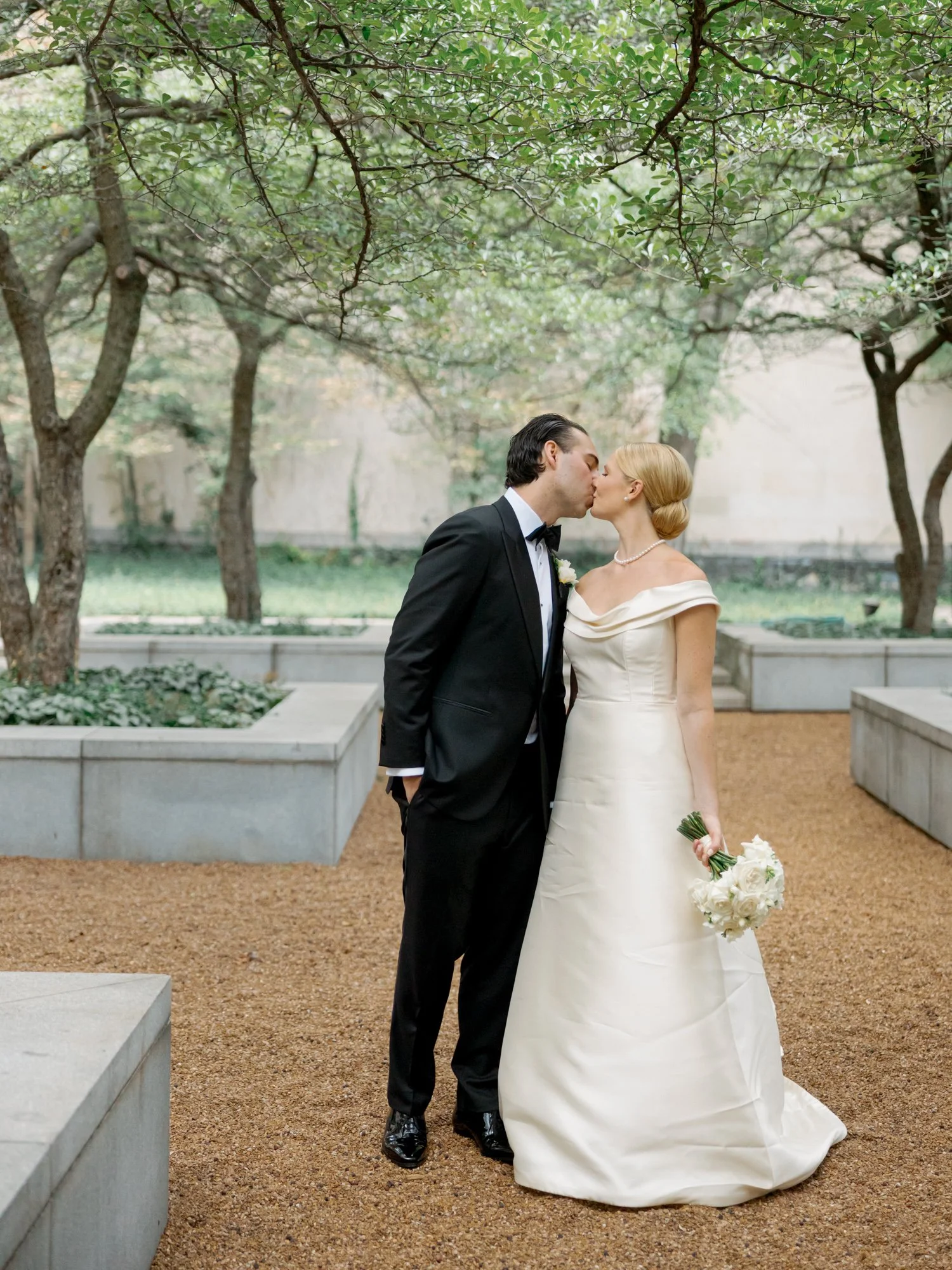Bride and groom share a soft kiss in an outdoor garden courtyard under bare-branched trees, her white bouquet at her side, completely still together.
