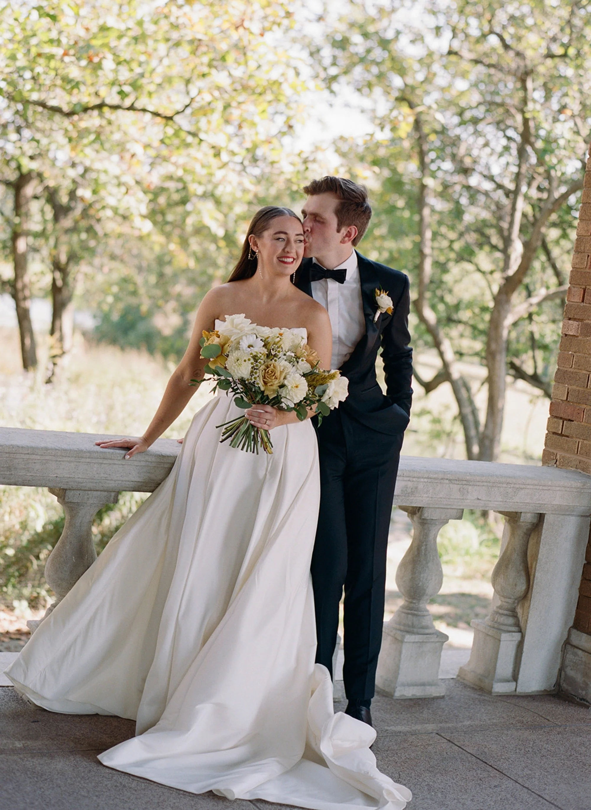 The groom plants a kiss on his bride's cheek as she leans against the stone balustrade of Columbus Park Refectory — golden autumn trees soft behind them, her bouquet at her side
