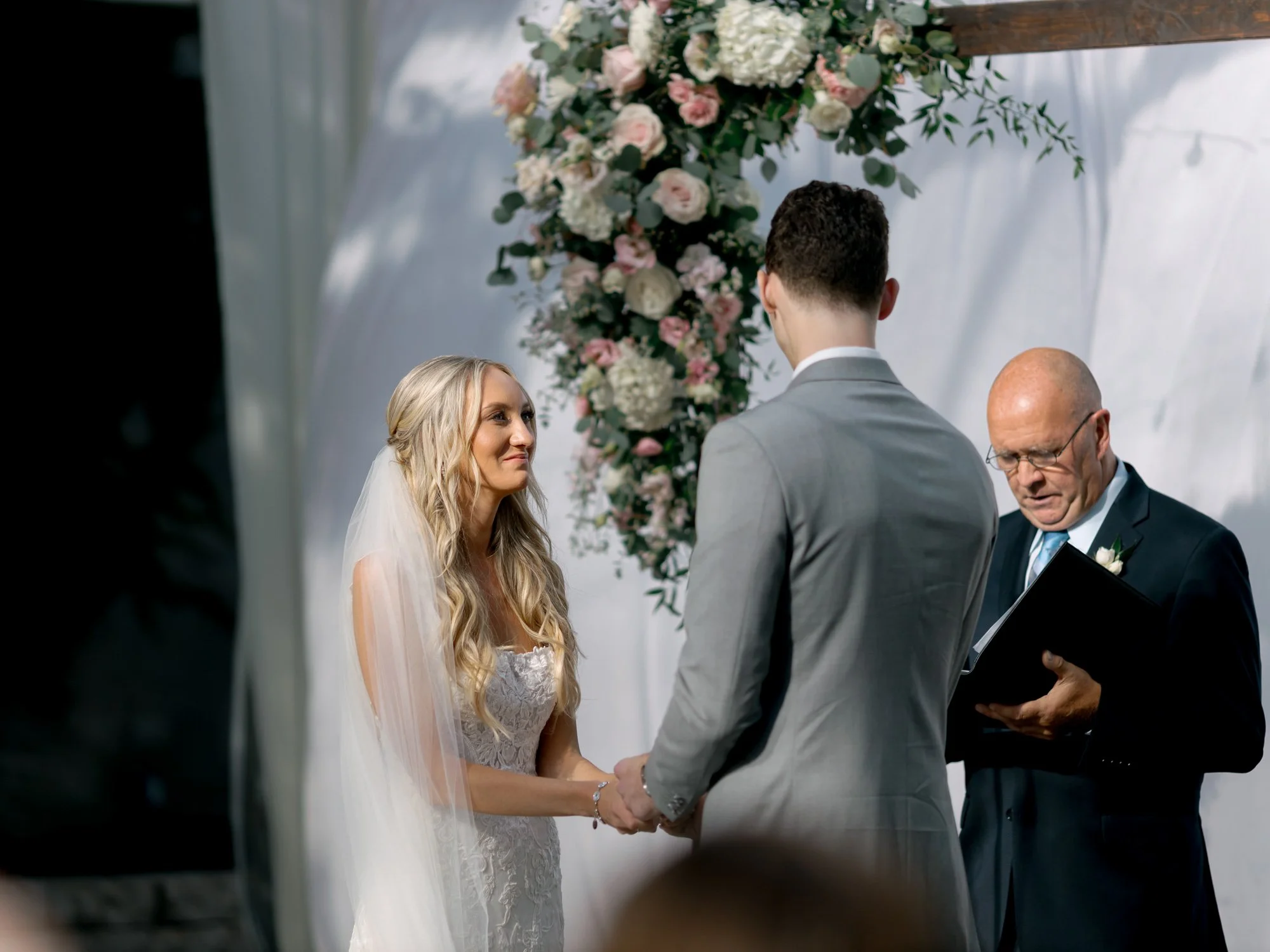 The bride beams up at her groom as they clasp hands during the ceremony vows, the blush floral arch and soft white draping of the venue behind them.