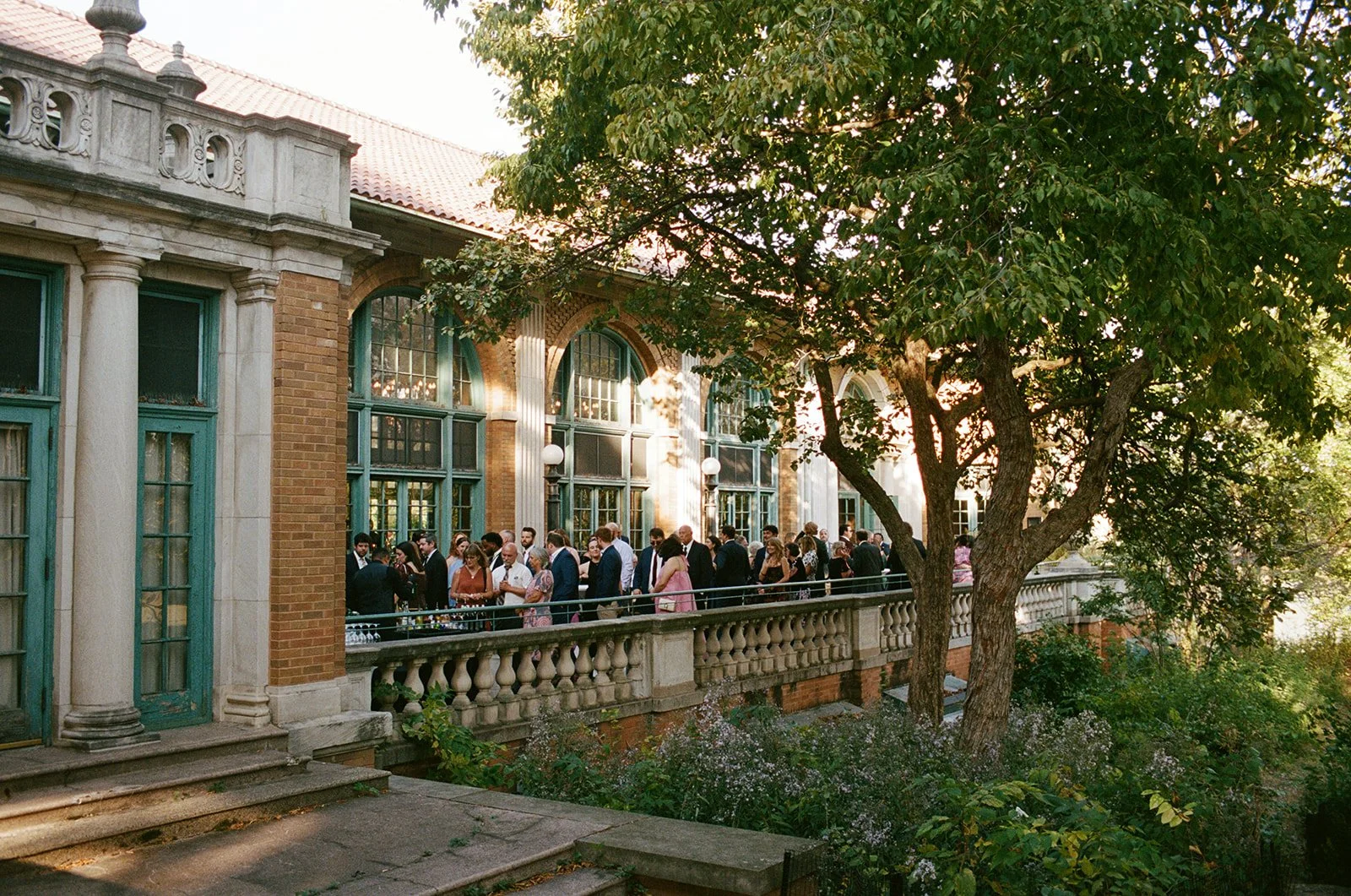 Wedding guests mingling on the sun-drenched stone terrace of Columbus Park Refectory during cocktail hour, the historic brick facade and teal arched windows glowing in golden light — Columbus Park Refectory wedding, Chicago film wedding photographer.