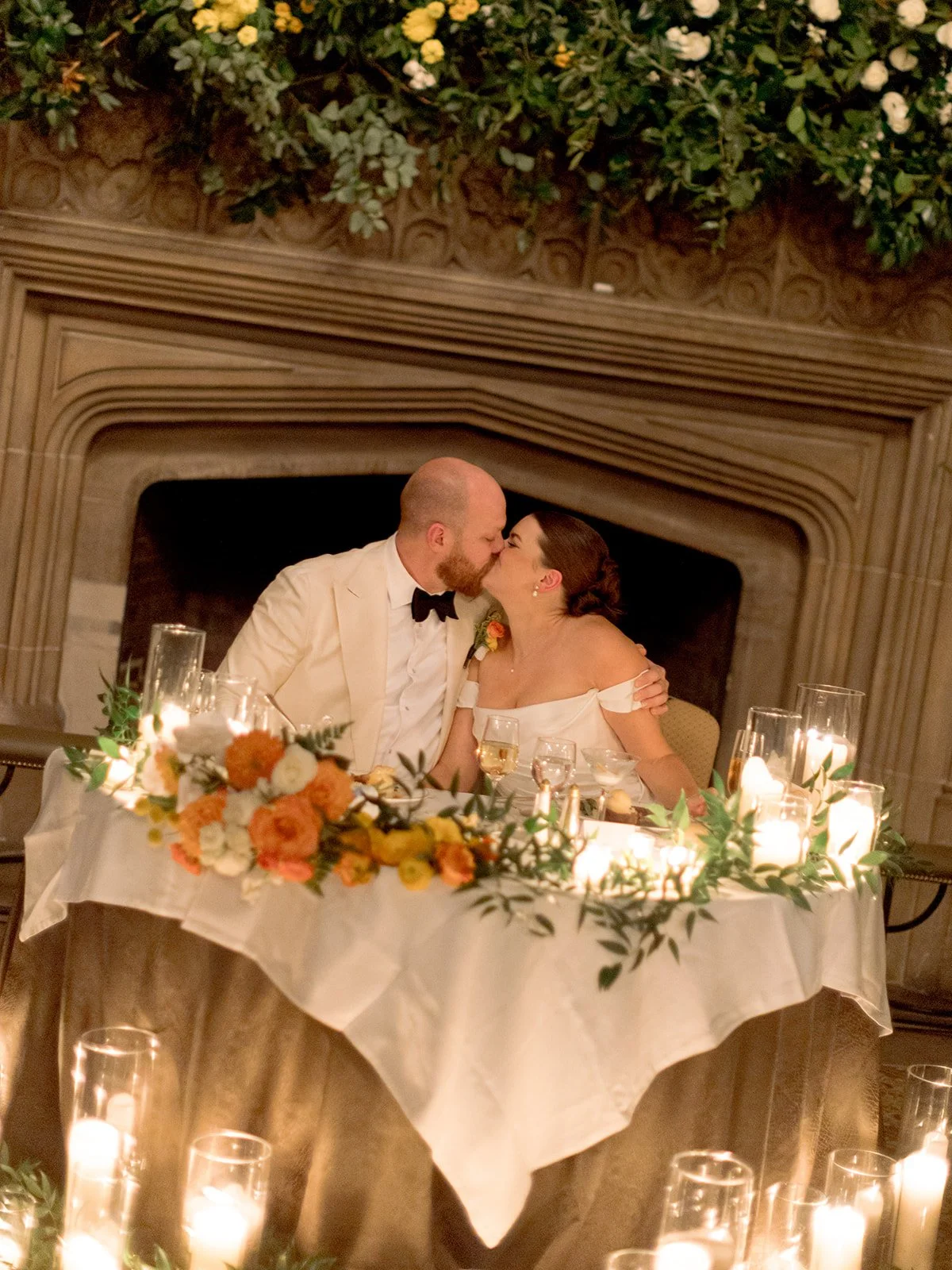 The couple shares a quiet kiss at their sweetheart table, the University Club Chicago's stone fireplace behind them and a lush greenery-and-floral installation cascading above, candles glowing on all sides.