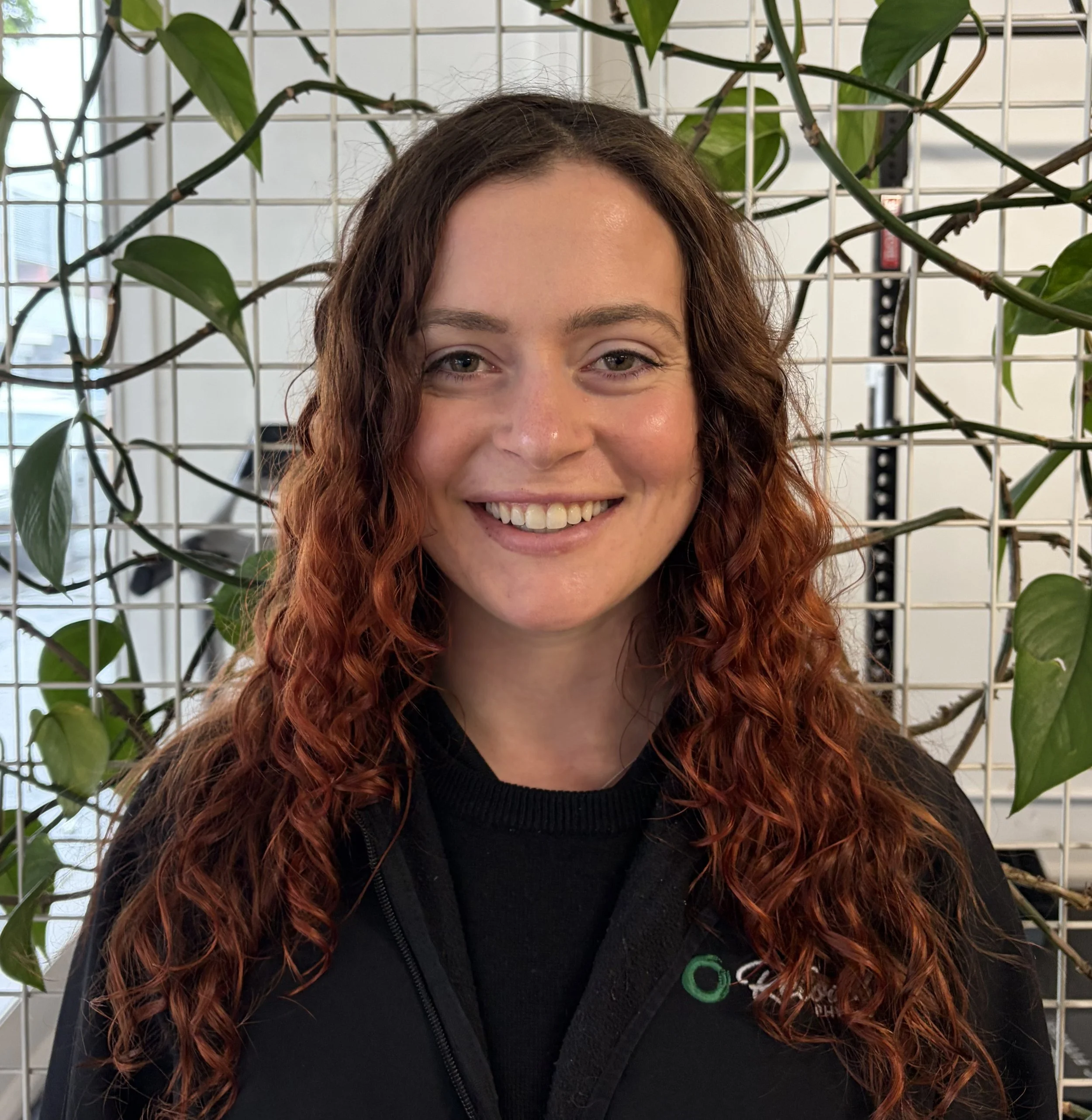 A woman with curly red hair smiling in front of a leafy background and a grid metal structure.