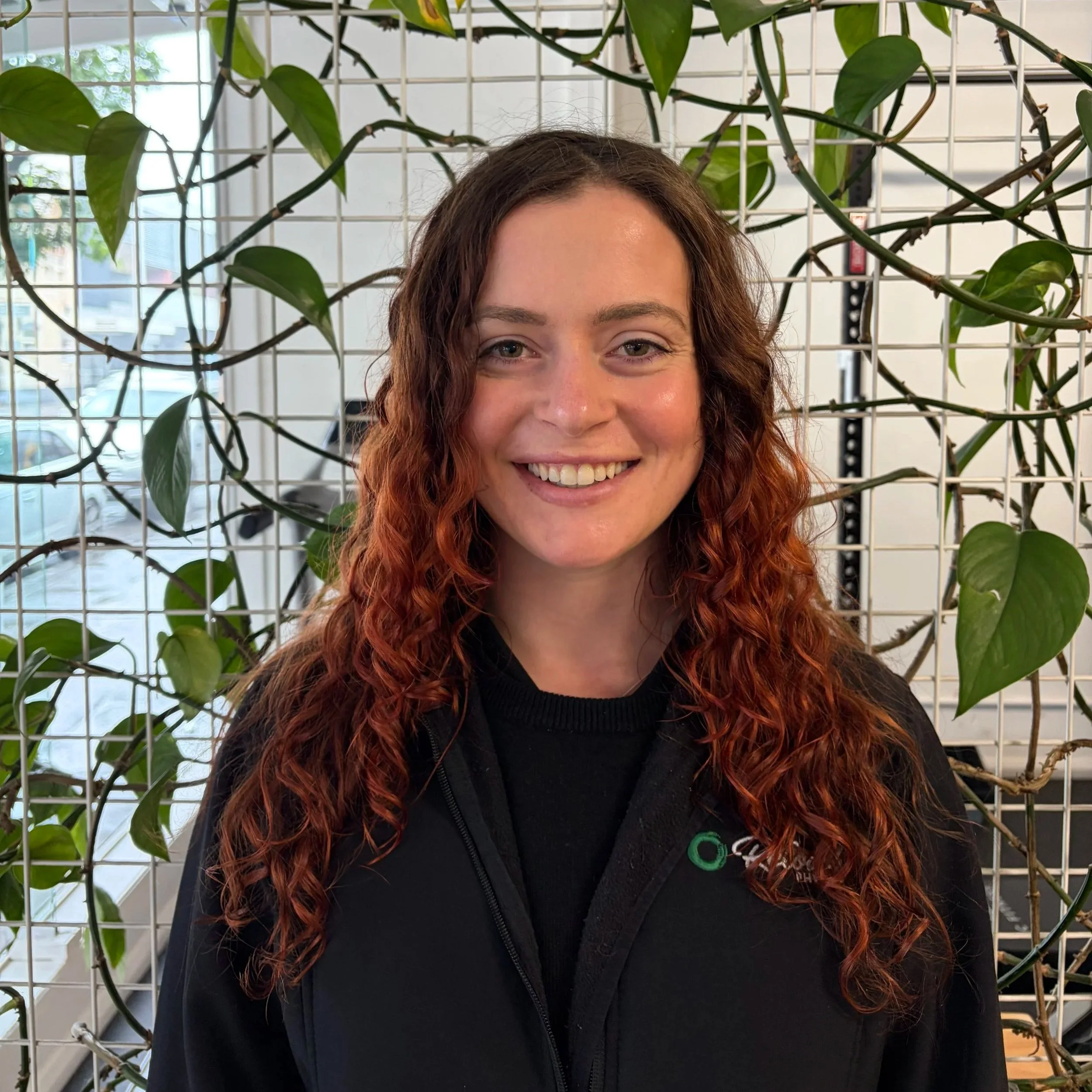 A young woman with curly hair smiling in front of green leafy plants.