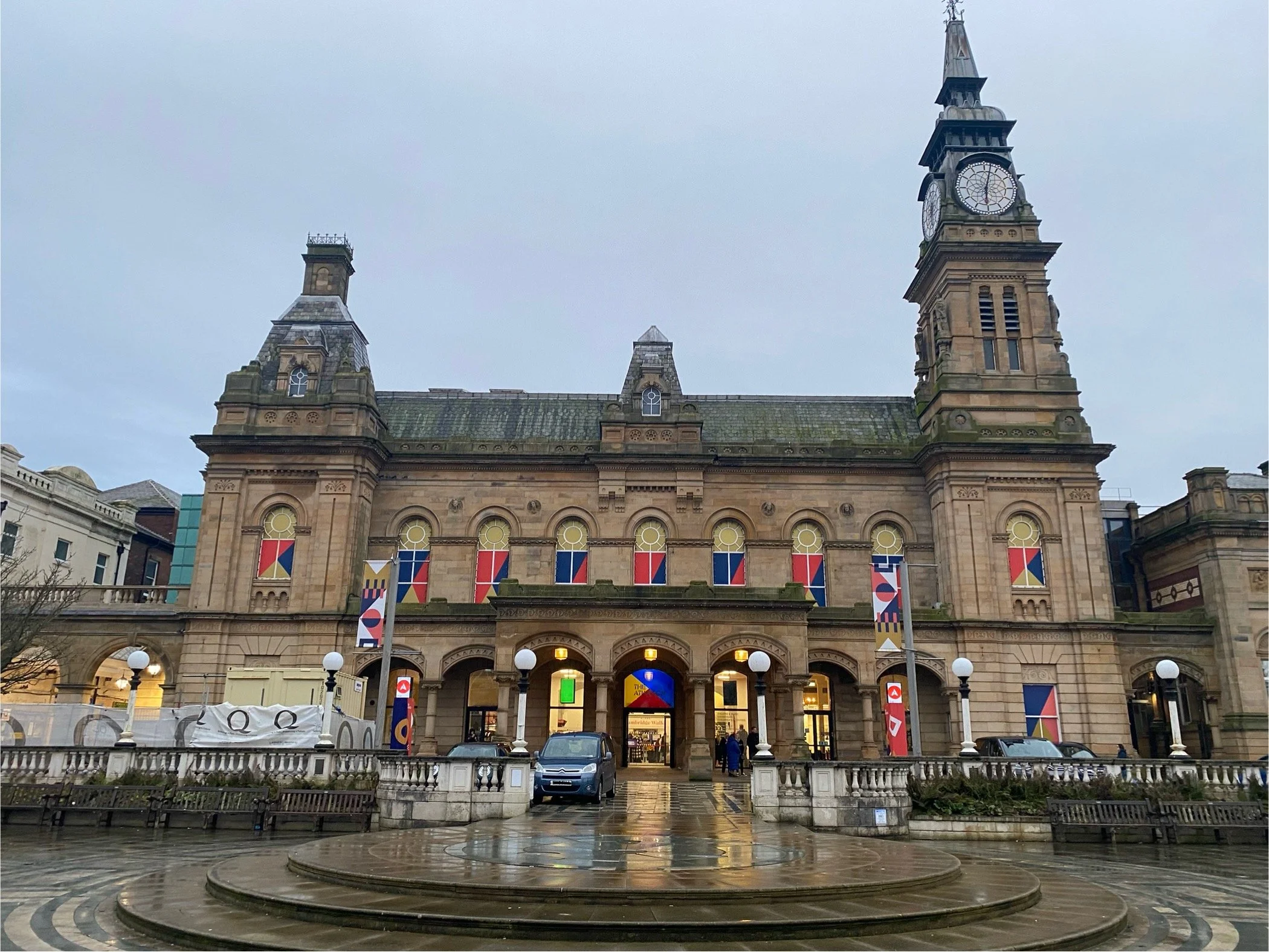 Front view of a historic stone building with two clock towers, colorful banners, and a set of steps leading to the entrance, with a wet pavement reflecting the building, on a cloudy day.