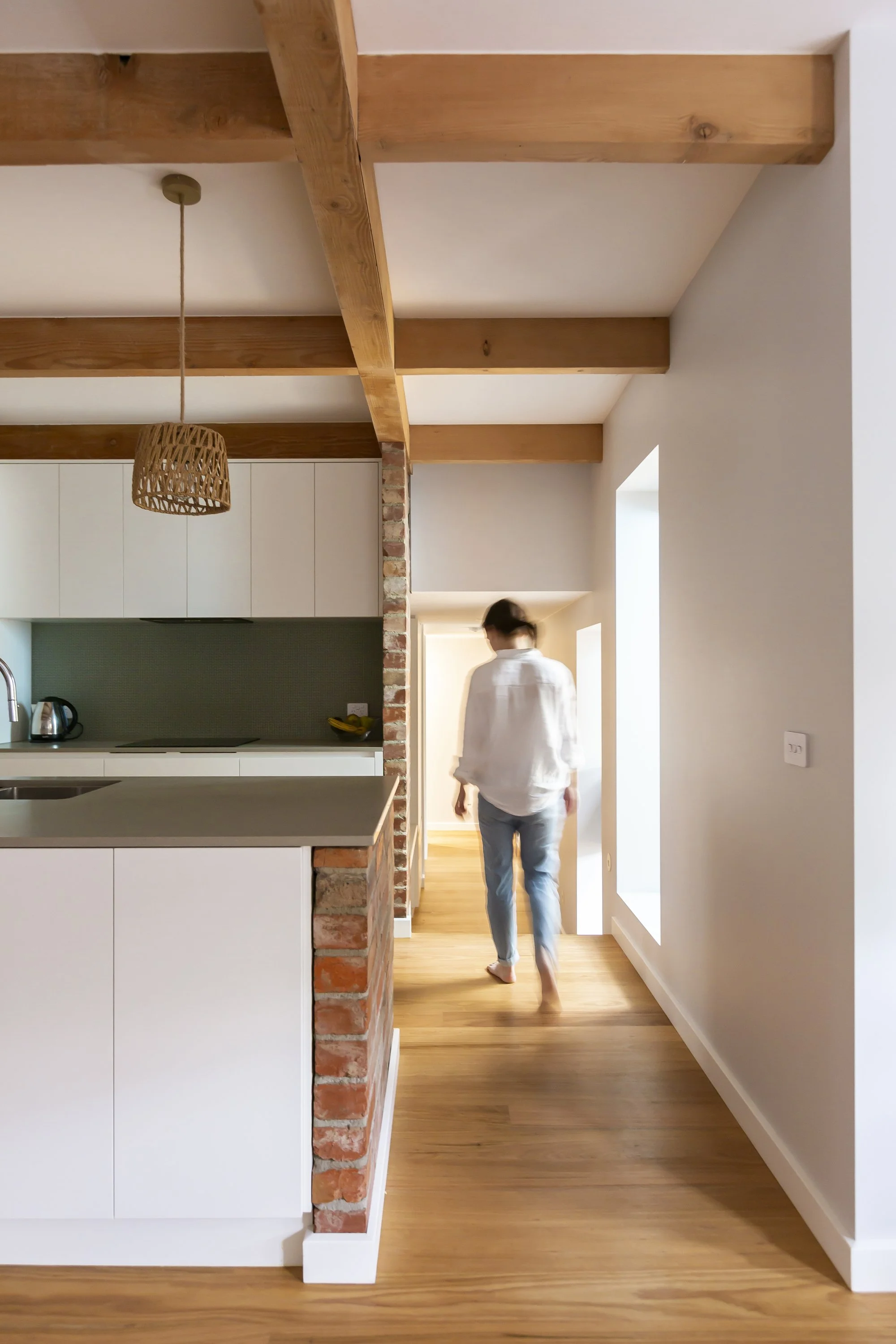 Interior of a modern home with exposed wooden beams on the ceiling, a white kitchen with a brick divider, and a woman walking barefoot towards a brightly lit hallway.