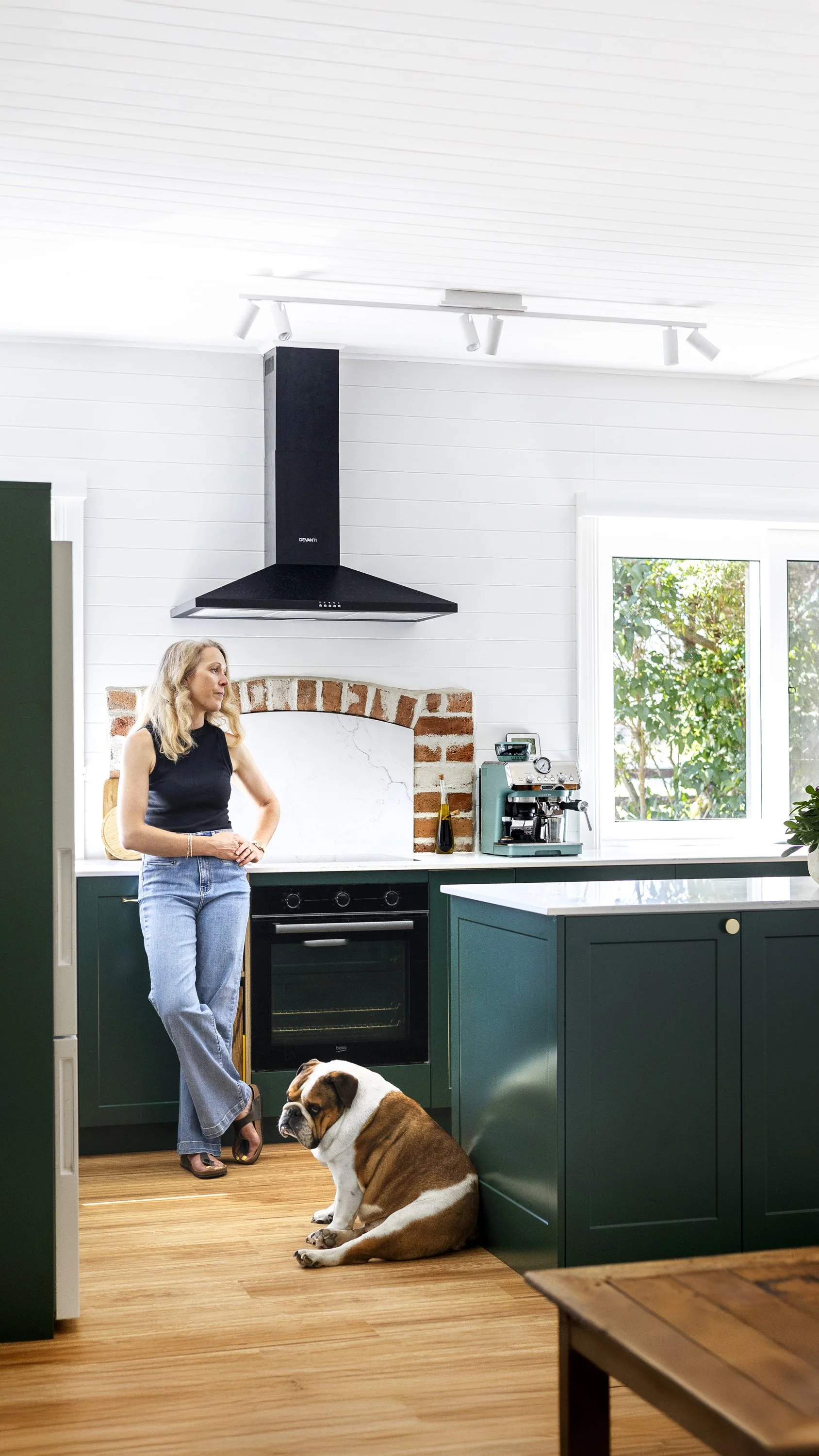 A woman standing in a modern green kitchen with a dog sitting on wooden floor near her, a window showing greenery outside, and various kitchen appliances.