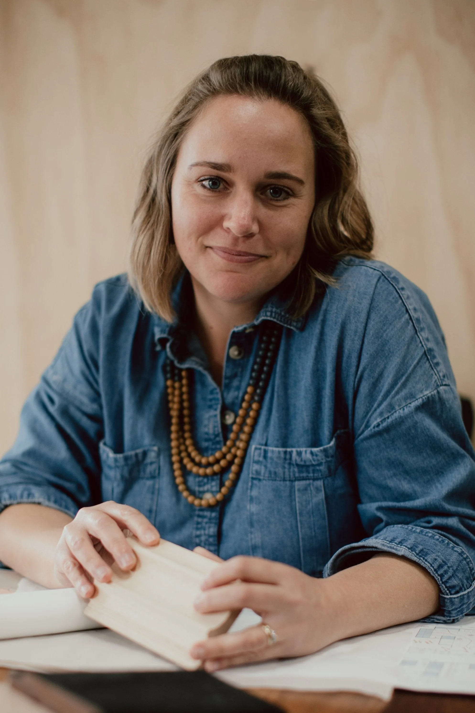 A woman with shoulder-length brown hair, blue eyes, and light skin, wearing a denim shirt and a beaded necklace, sitting at a table with an open book.
