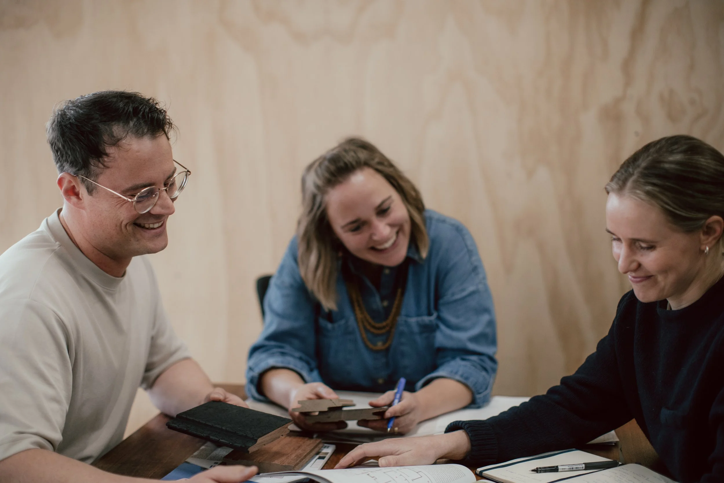 Three people sitting around a table, smiling and discussing something, with color swatches and notebooks on the table.