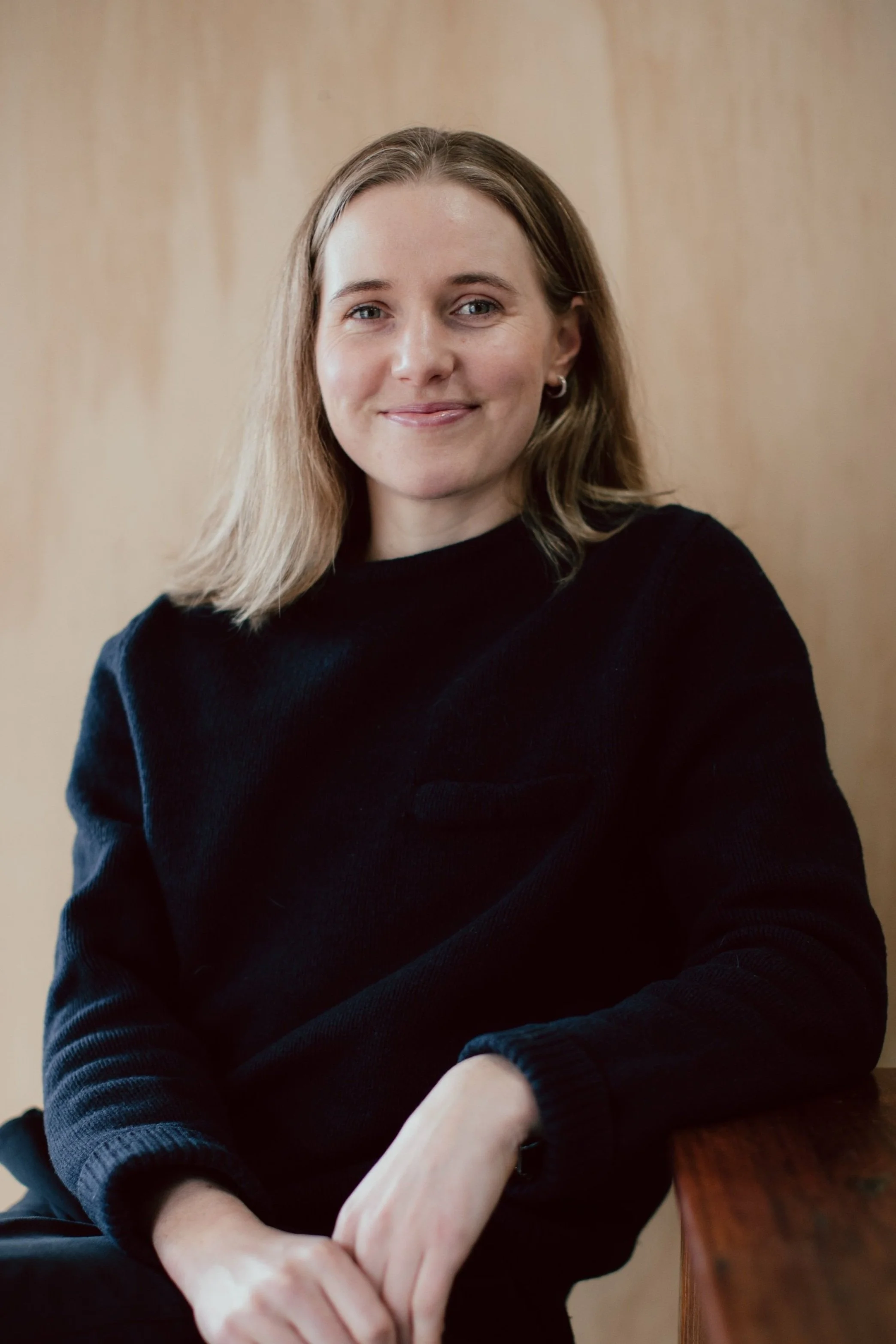 A woman with shoulder-length light brown hair smiling, wearing a black sweater, sitting against a wooden background.