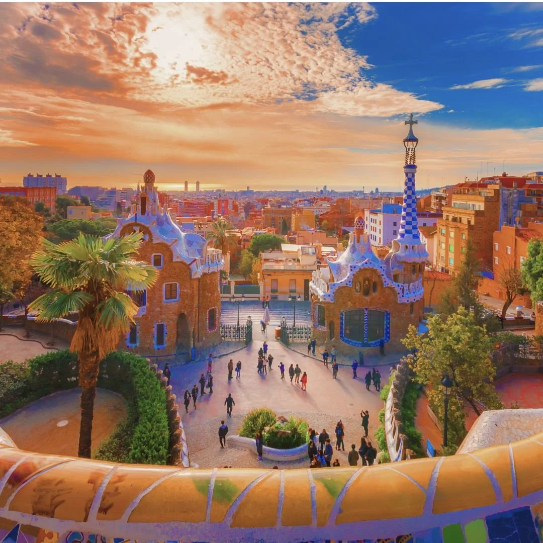 Colorful sunset view of Park Güell in Barcelona, Spain, featuring whimsical mosaic-tile buildings, a pathway with visitors, and a city skyline in the background.