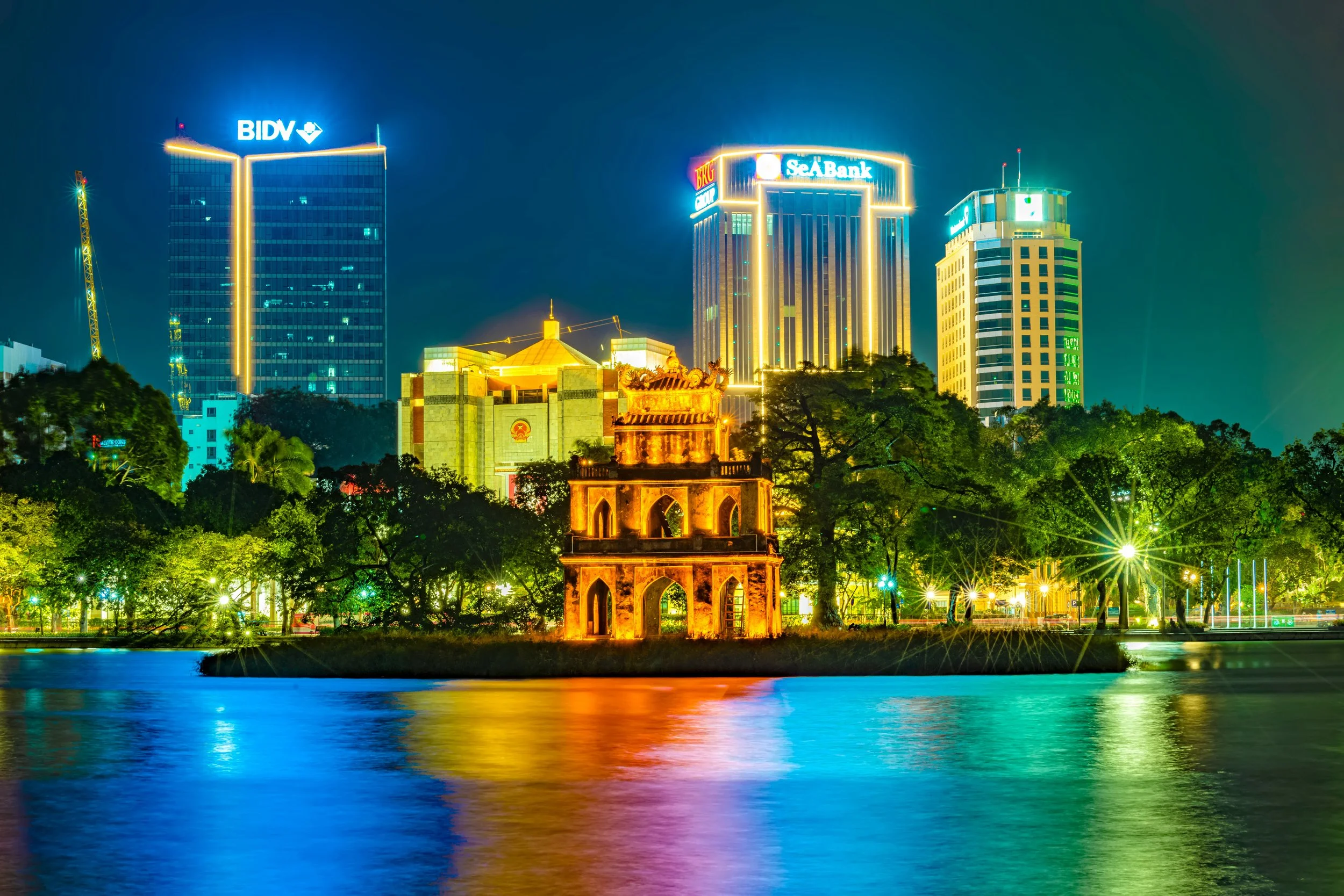 Night view of a city skyline with tall illuminated buildings, a historic tower in the foreground, and colorful reflections in the water.