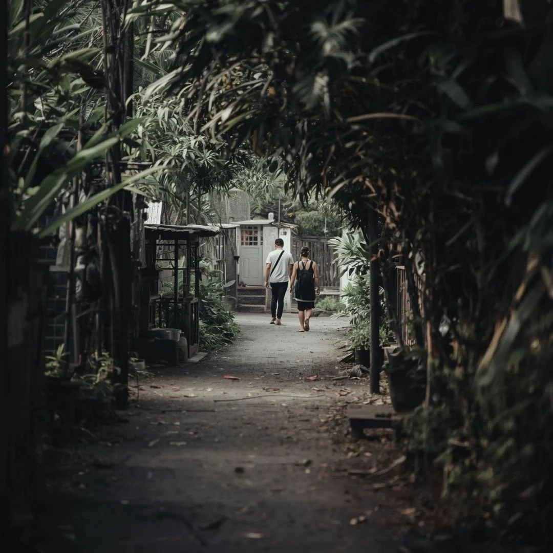 Couple walking through a quiet village pathway in Bali surrounded by greenery, captured in a natural everyday moment