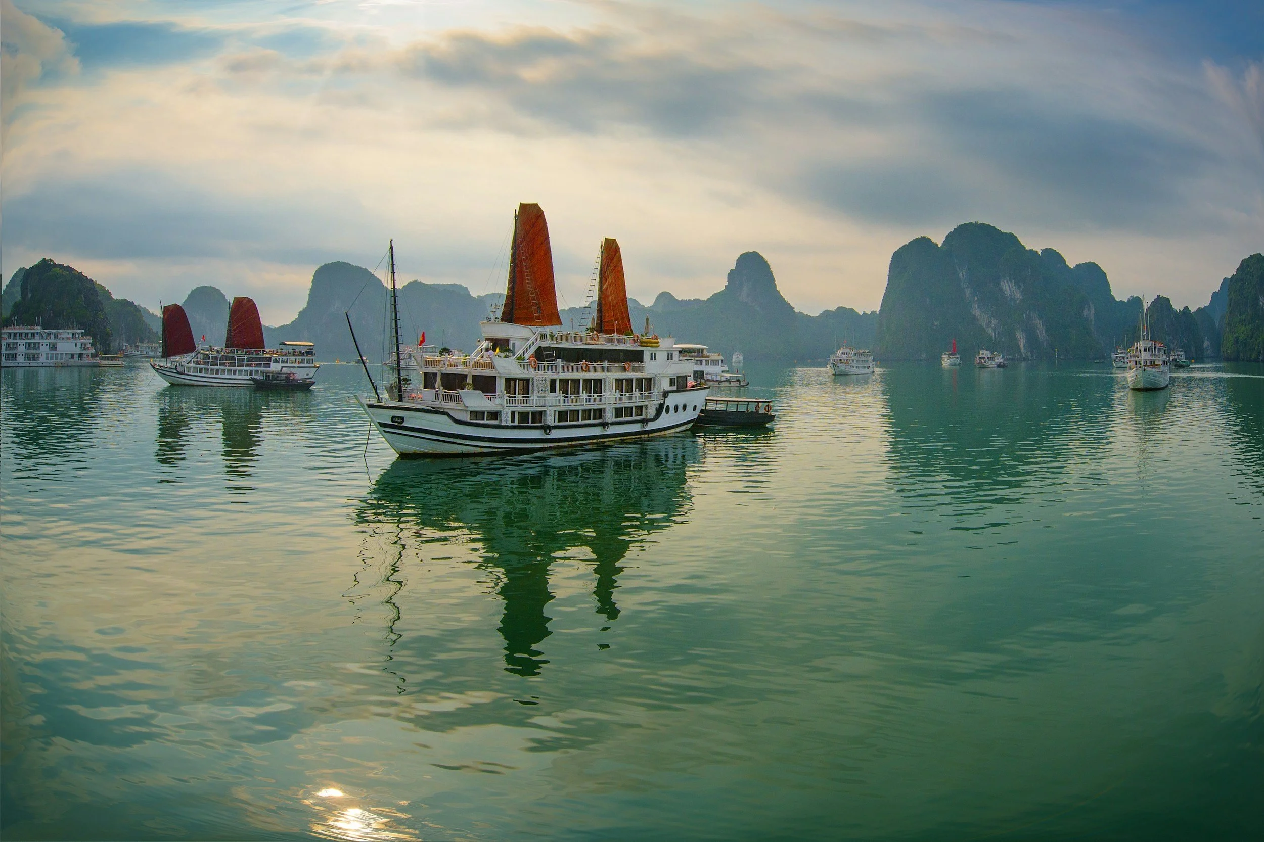 Several traditional Asian-style boats with brown sails floating on calm water, set against limestone karst islands under a cloudy sky.