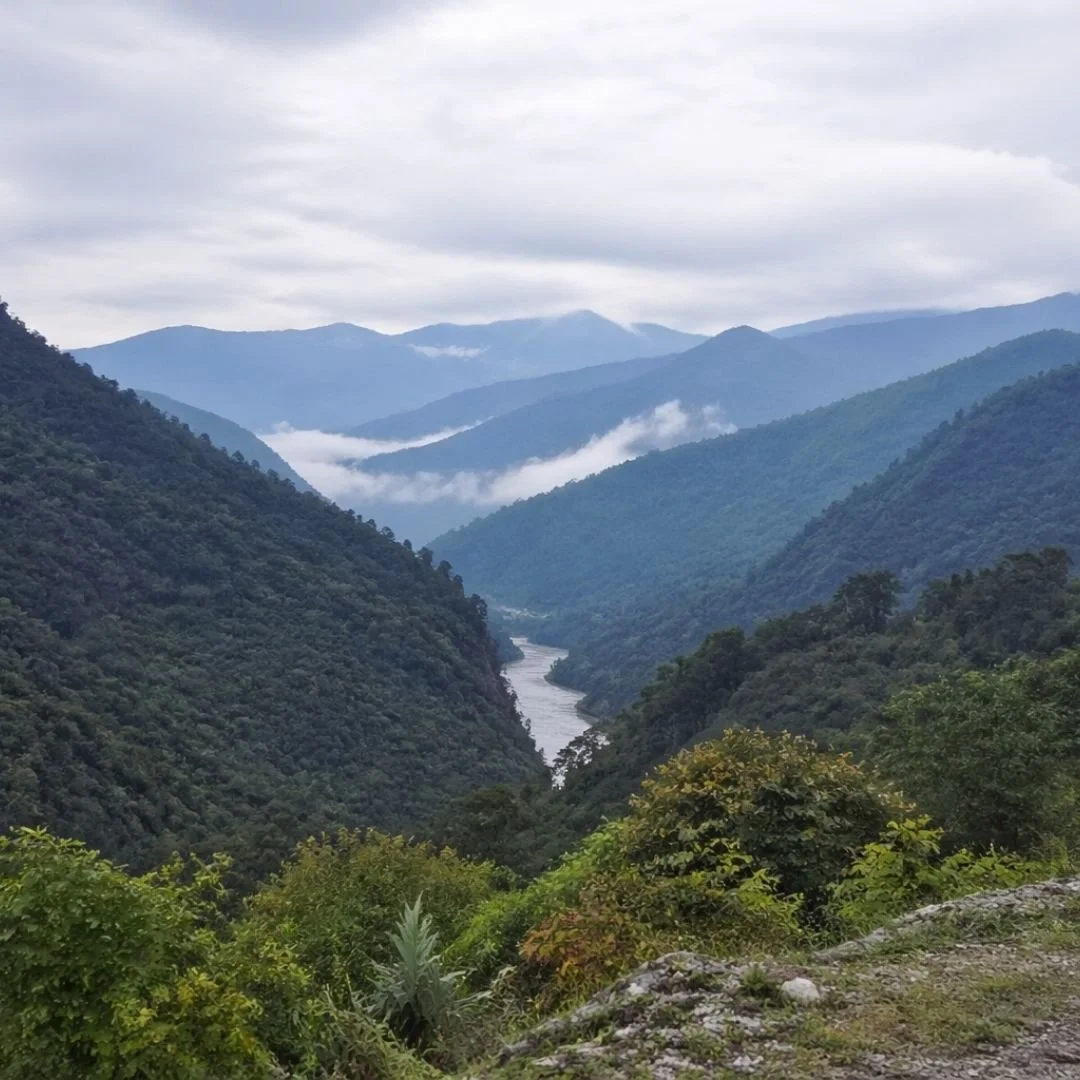 Wide-angle view of a misty mountain valley in Arunachal Pradesh with layered forested hills and a pale river flowing far below under soft overcast light