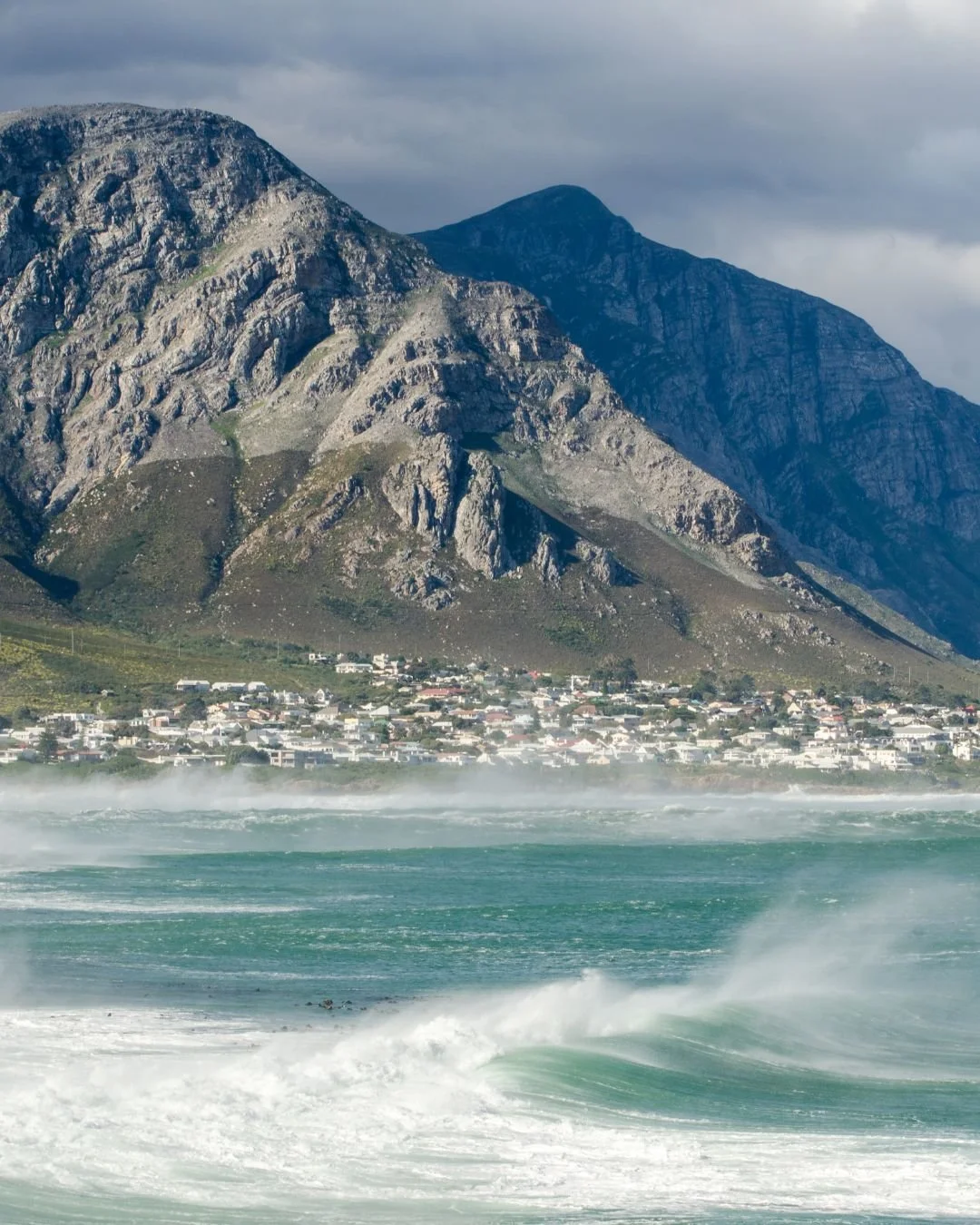 A coastal town at the base of rugged mountains, with ocean waves and mist in the foreground.