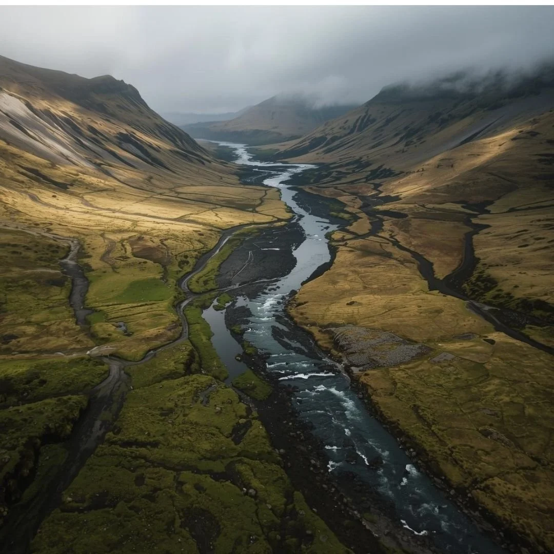 An aerial view of a winding river cutting through an expansive Icelandic valley, with mossy terrain, layered mountains, and low clouds creating a sense of vast, untouched wilderness.