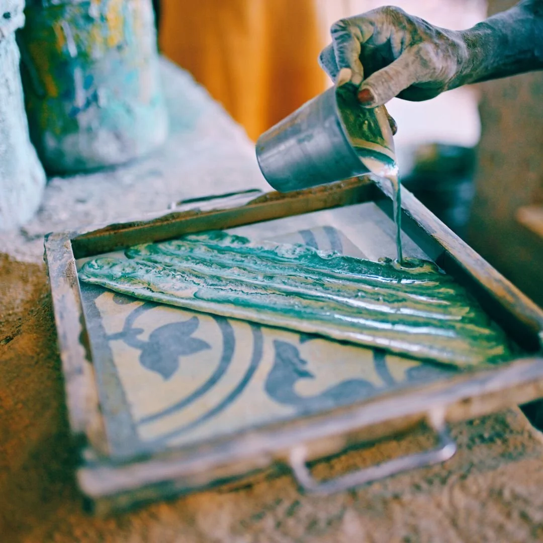 Person pouring green and white marbled liquid onto a patterned tray as part of a tile-making process.