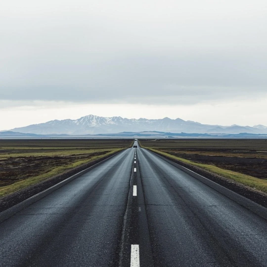 A long, empty road stretching straight through a vast open landscape, leading toward distant mountains under a wide, overcast sky, conveying quiet isolation and scale.